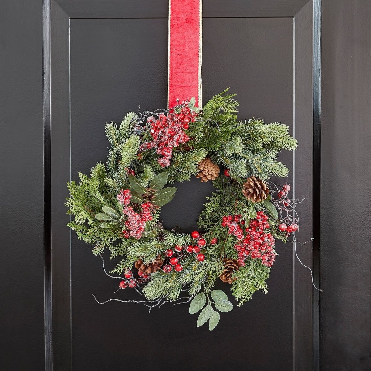 A frosted Christmas wreath with pinecones and red berries hanging on a black door by a red ribbon.