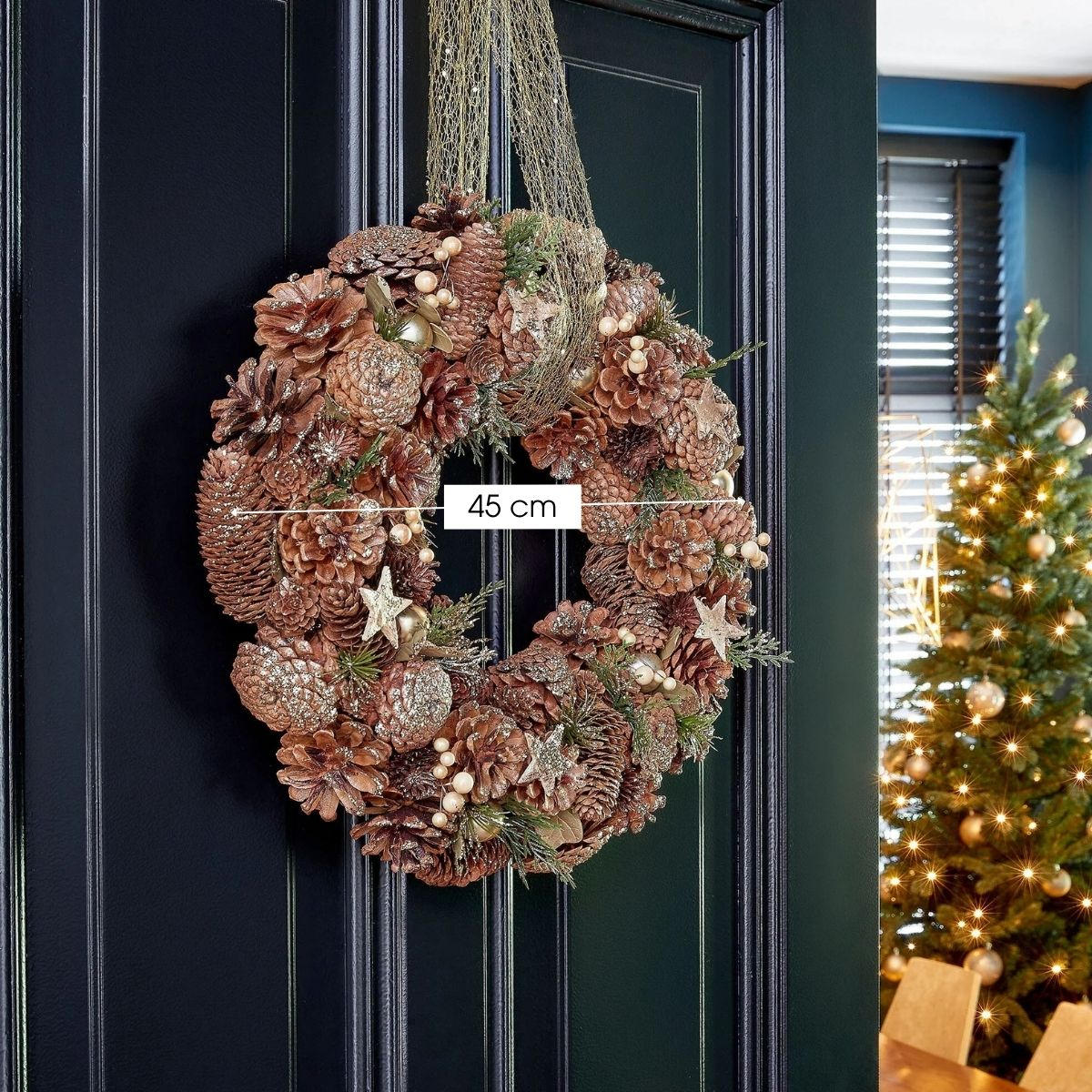 A 45 cm pinecone Christmas wreath hangs on a black door, with a Christmas tree in the background.
