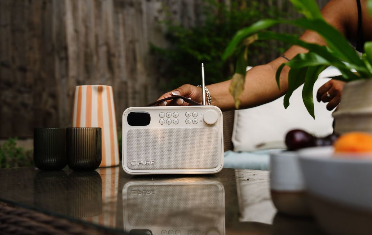Una radio Pure blanca se encuentra en una mesa al aire libre con plantas verdes y una jarra a rayas al fondo.
