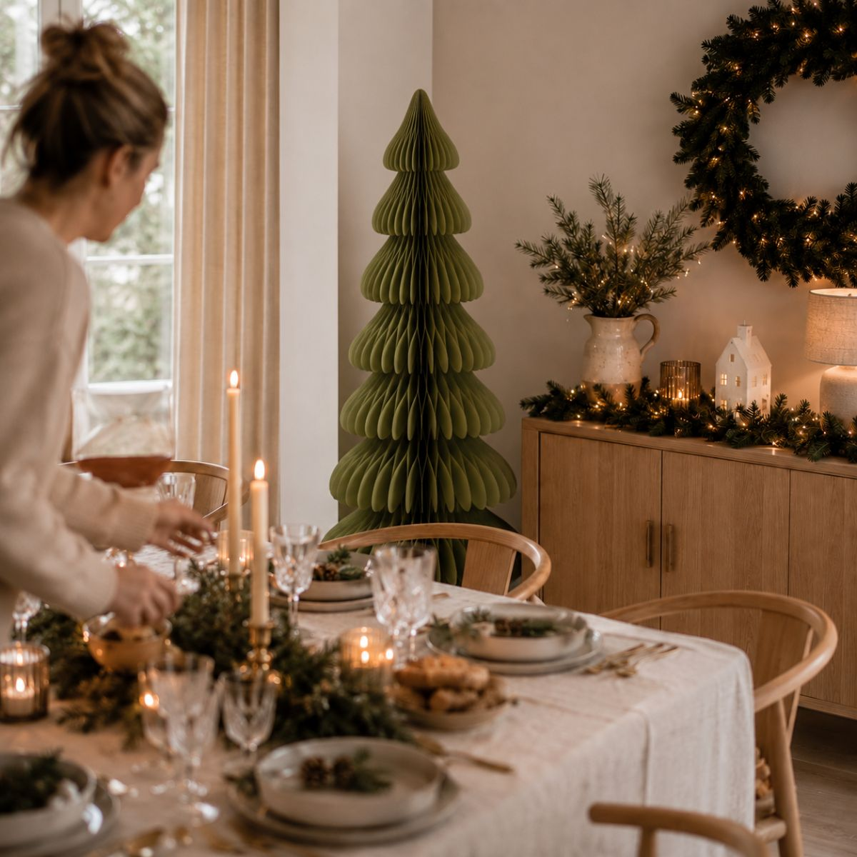 A woman sets a festive dining table in a Christmas-decorated room featuring a green paper tree.