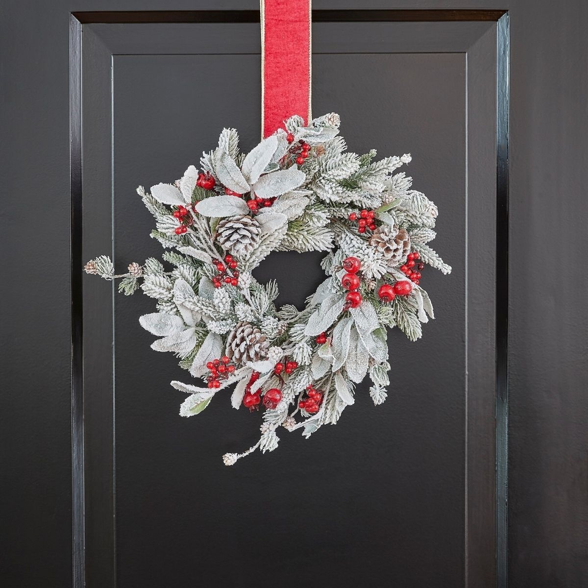 A snowy Christmas wreath with red berries and pinecones hangs on a dark door by a red ribbon.