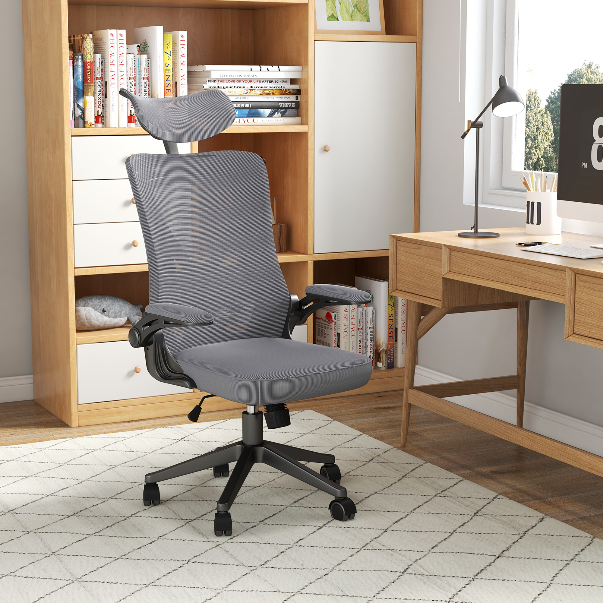 Una sedia da ufficio ergonomica grigia si trova davanti a una libreria in legno e una scrivania.