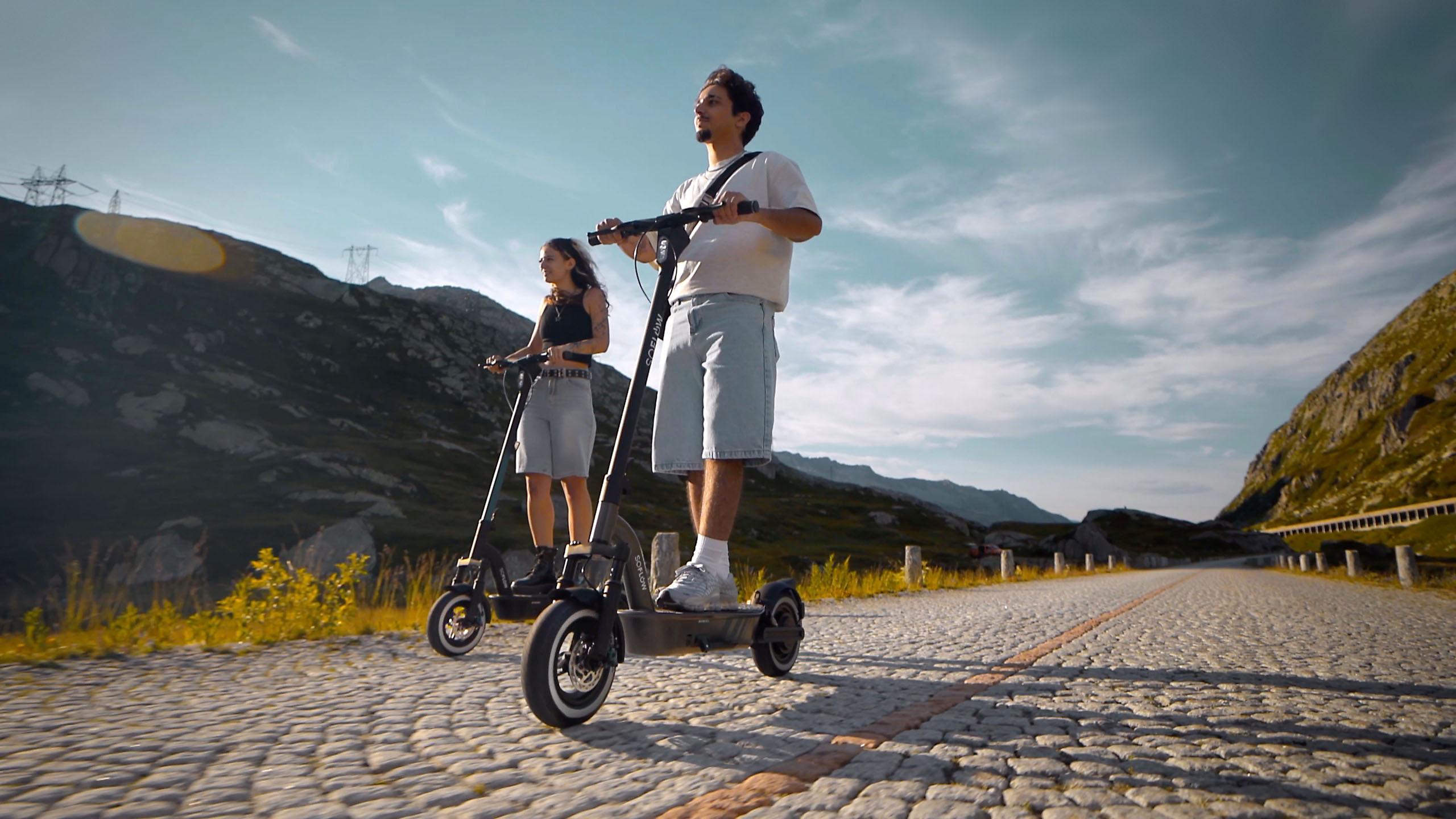 Ein Mann und eine Frau fahren E-Scooter auf einer Kopfsteinpflasterstraße in den Bergen unter blauem Himmel.