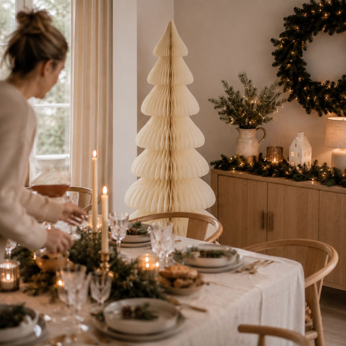 Une femme dresse une table de salle à manger aux couleurs de Noël avec des bougies et un grand sapin en papier.