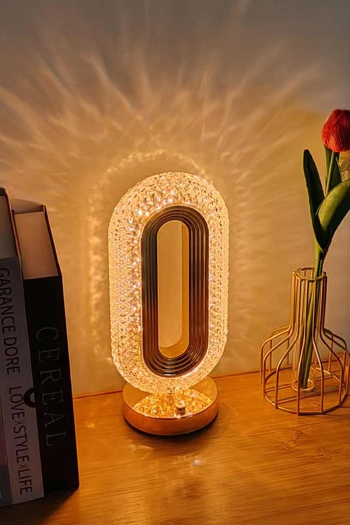 Işıklı oval kristal lamba, ahşap bir yüzey üzerinde kitapların ve altın tel vazodaki kırmızı lalenin yanında duruyor.
