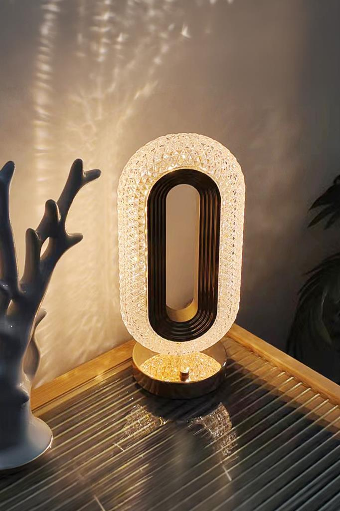 Koyu merkezli oval kristal masa lambası cam masada, duvara sıcak ışık yayıyor.