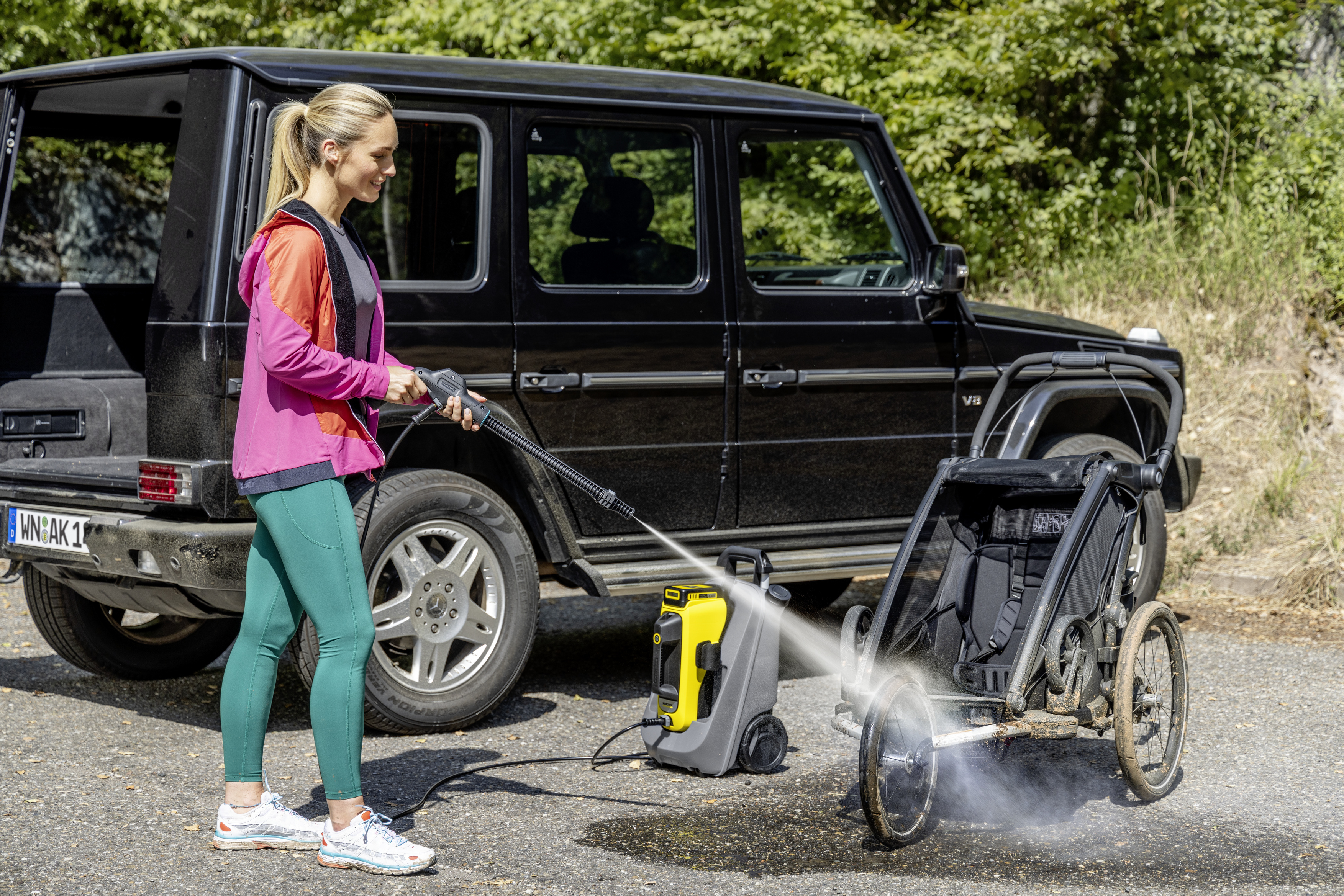 Eine Frau wäscht einen schmutzigen Kinderwagen mit einem Kärcher-Hochdruckreiniger vor einem schwarzen Auto.