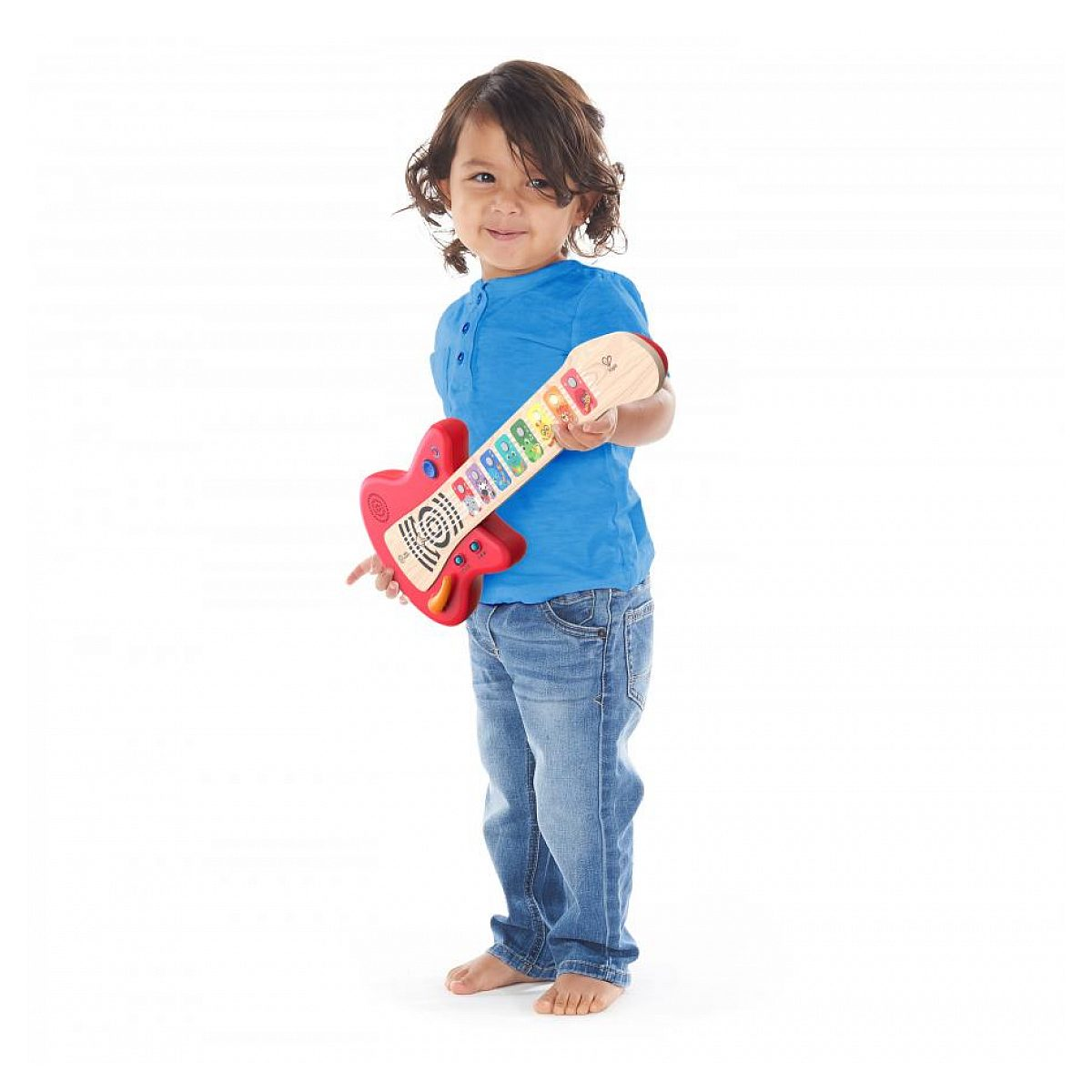 Un niño pequeño con camisa azul y jeans sonríe mientras sostiene una guitarra de juguete roja y de madera.