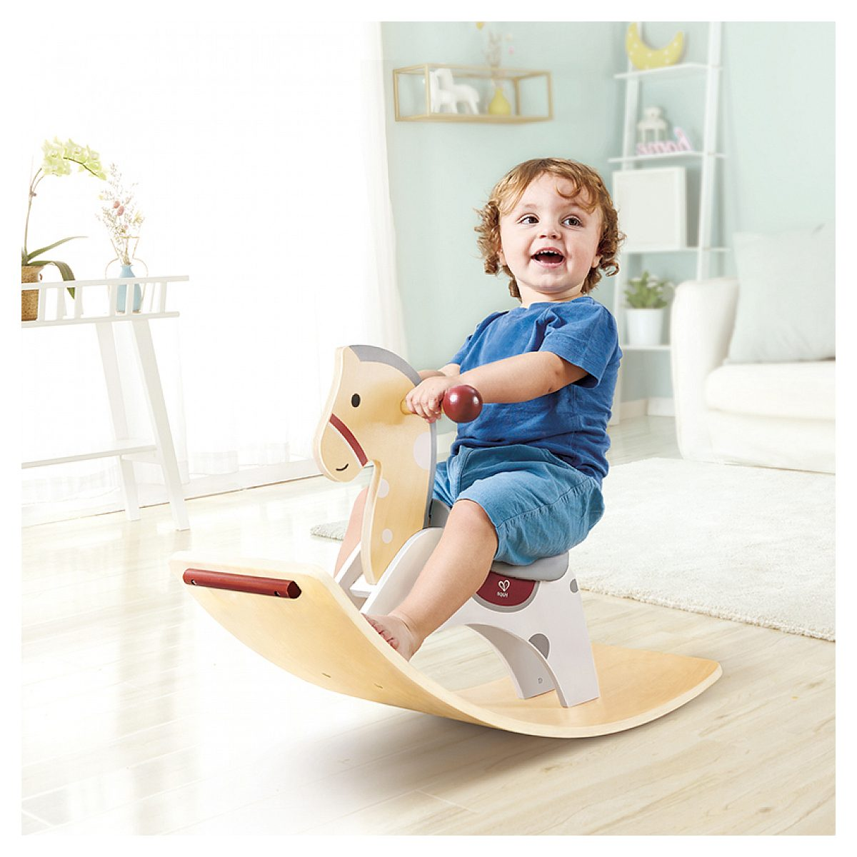 Kleinkind lacht und reitet auf einem hölzernen Schaukelpferd in einem hellen Kinderzimmer.