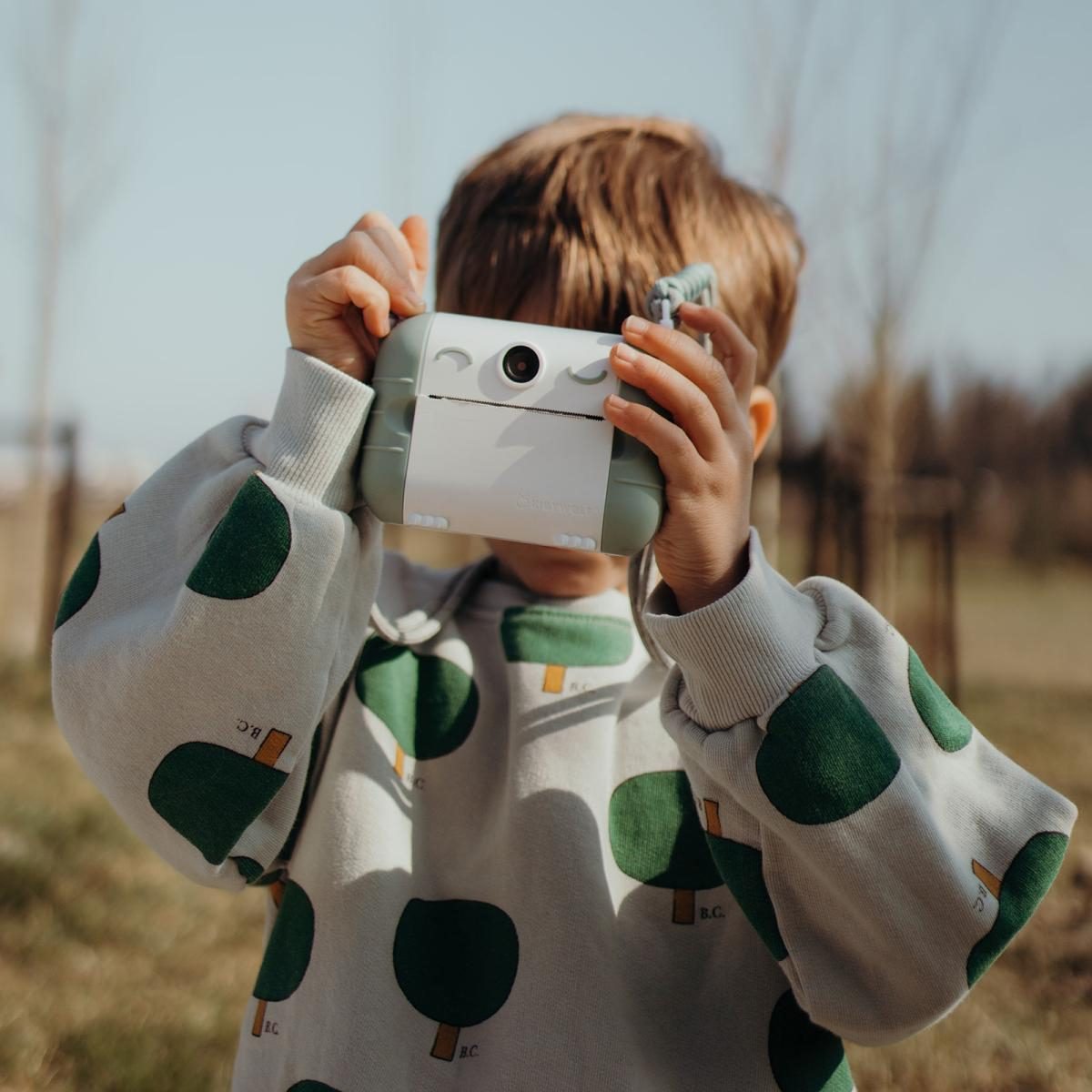 Ein Junge in einem grün gemusterten Pullover hält draußen eine weiß-grüne Spielzeugkamera vor das Gesicht.