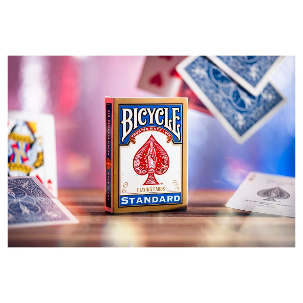 Una caja cerrada de cartas Bicycle Standard sobre una mesa de madera, rodeada de naipes dispersos.