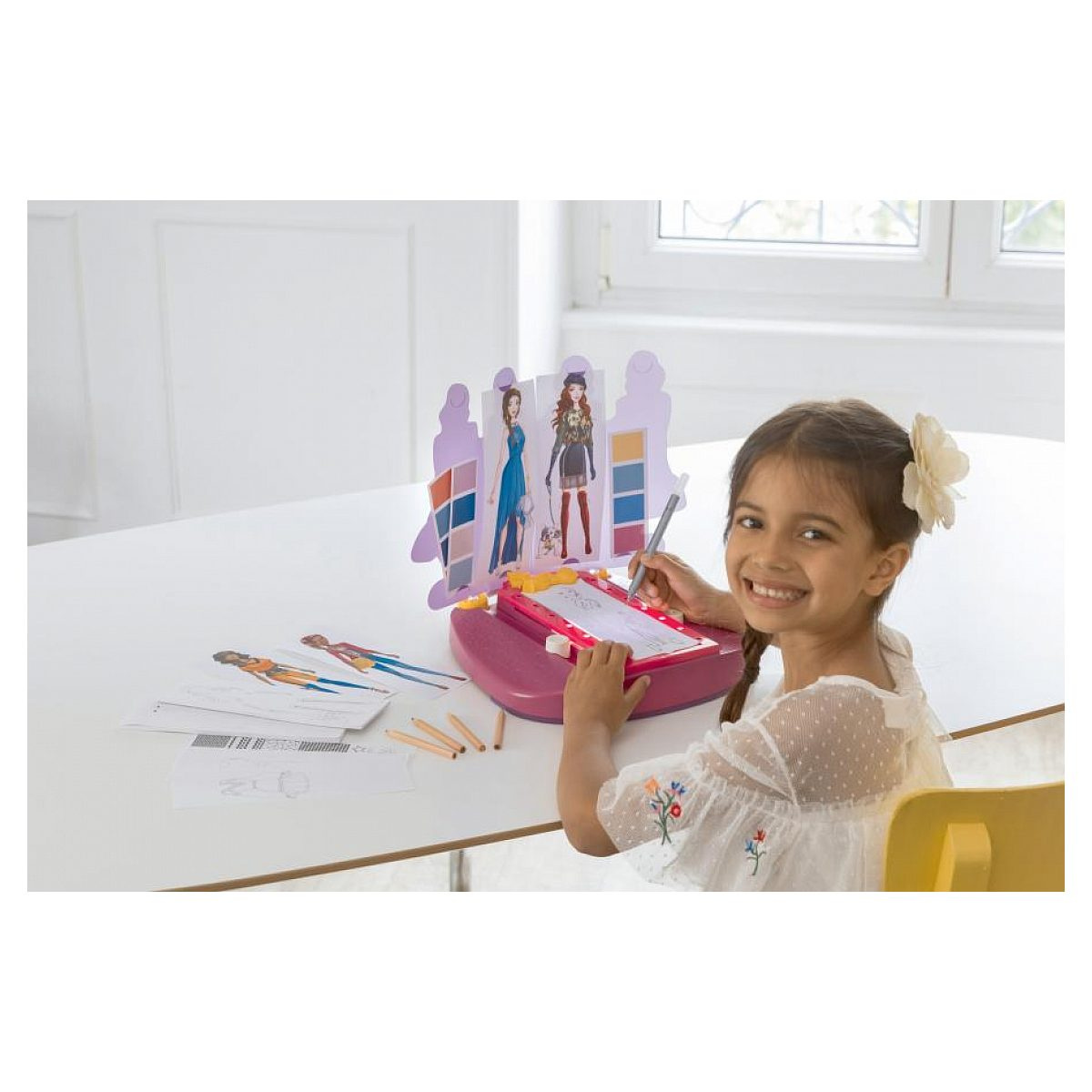 Ein lächelndes Mädchen sitzt am Tisch und zeichnet mit einem Modedesign-Set, umgeben von Skizzen.