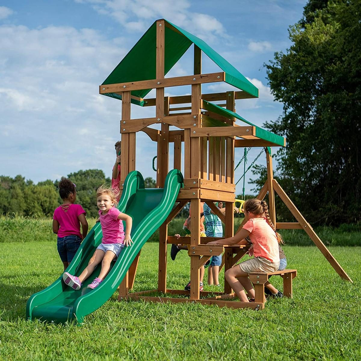 Kinder spielen auf einem Holzspielplatz mit grüner Rutsche und Dach auf einer Wiese.