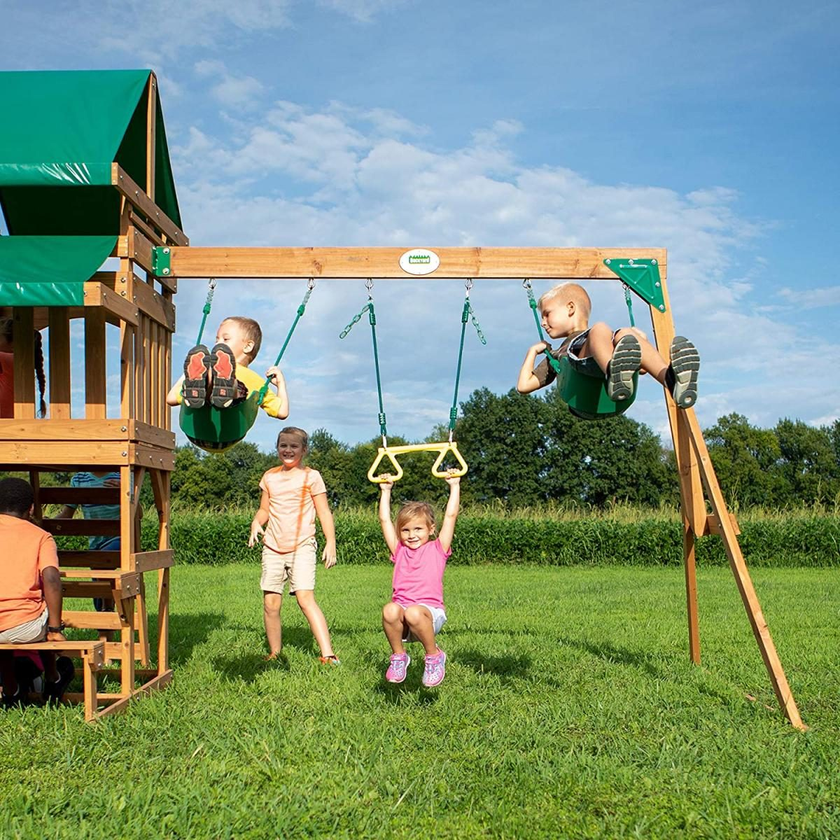 Kinder spielen auf einer Holzschaukel im grasbewachsenen Garten unter blauem Himmel.