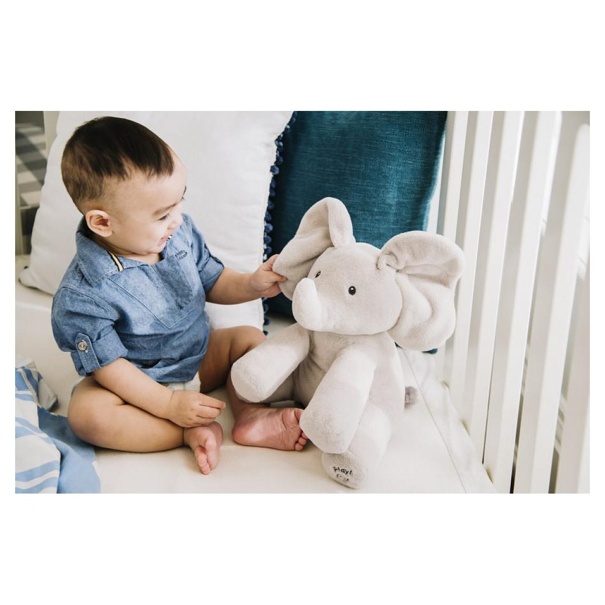 Ein Baby im blauen Hemd spielt in einem weißen Gitterbett mit einem grauen Kuschelelefant.