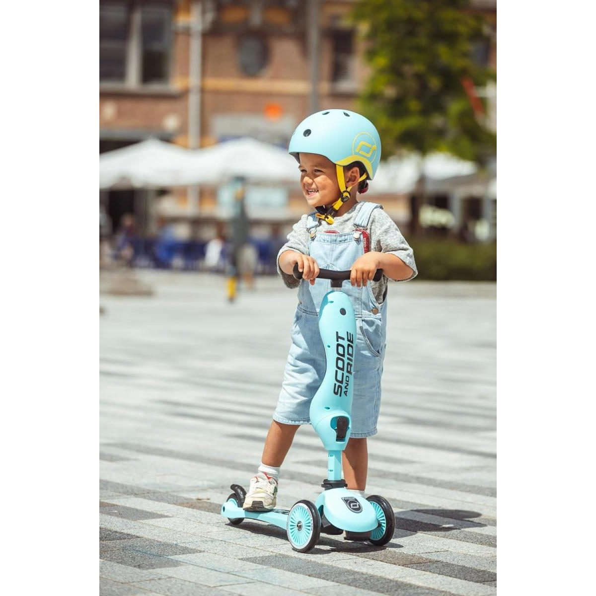 Un bambino sorridente con casco azzurro e salopette guida un monopattino azzurro all'aperto.