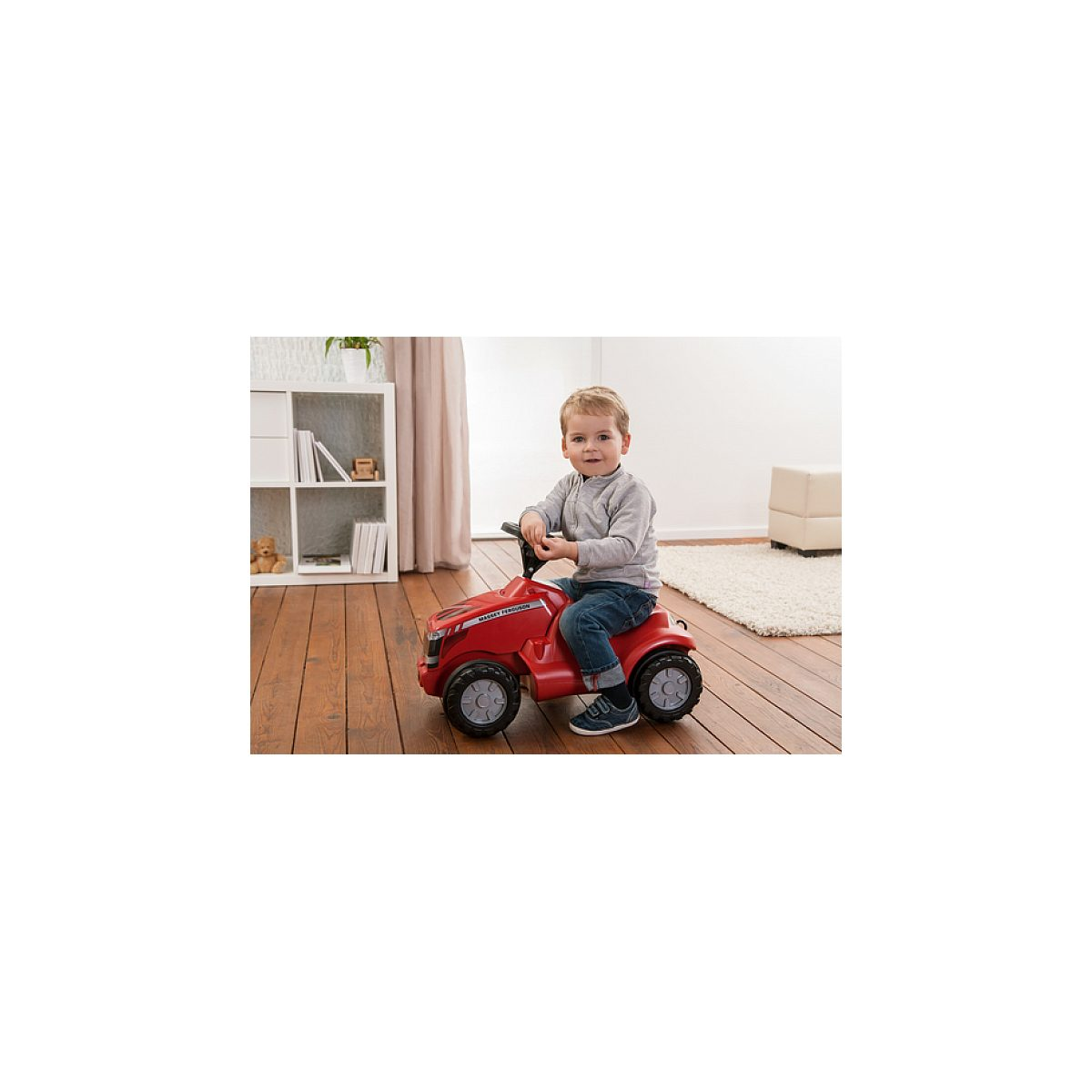 Kleinkind sitzt auf einem roten Spielzeugtraktor auf Holzboden in einem Zimmer.