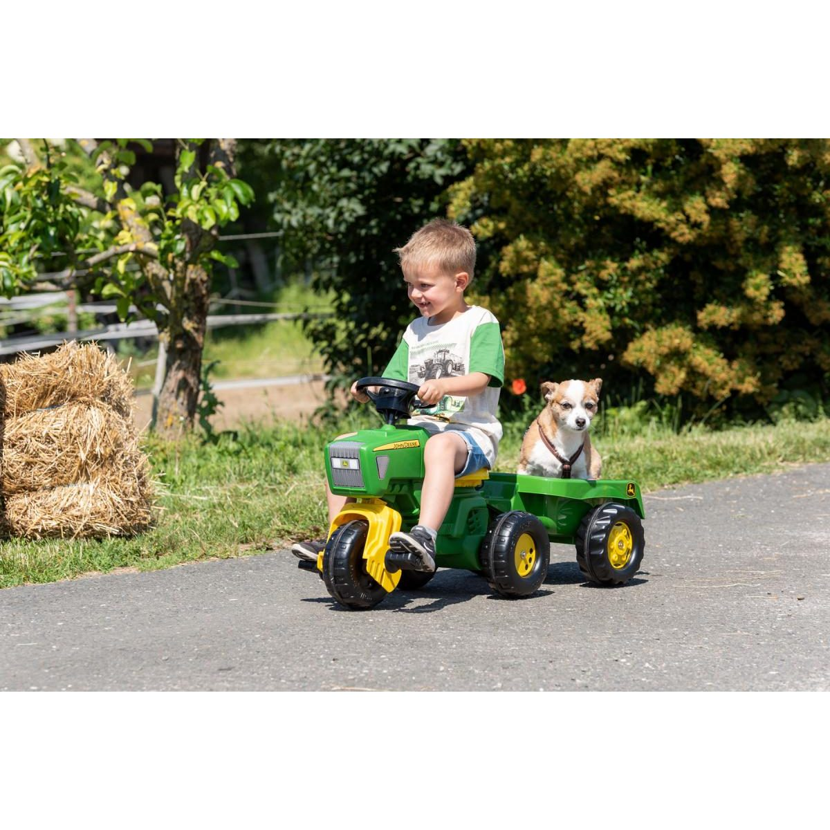 Ein Junge fährt einen grünen Spielzeugtraktor mit Hund im Anhänger auf einem Weg neben Heu.