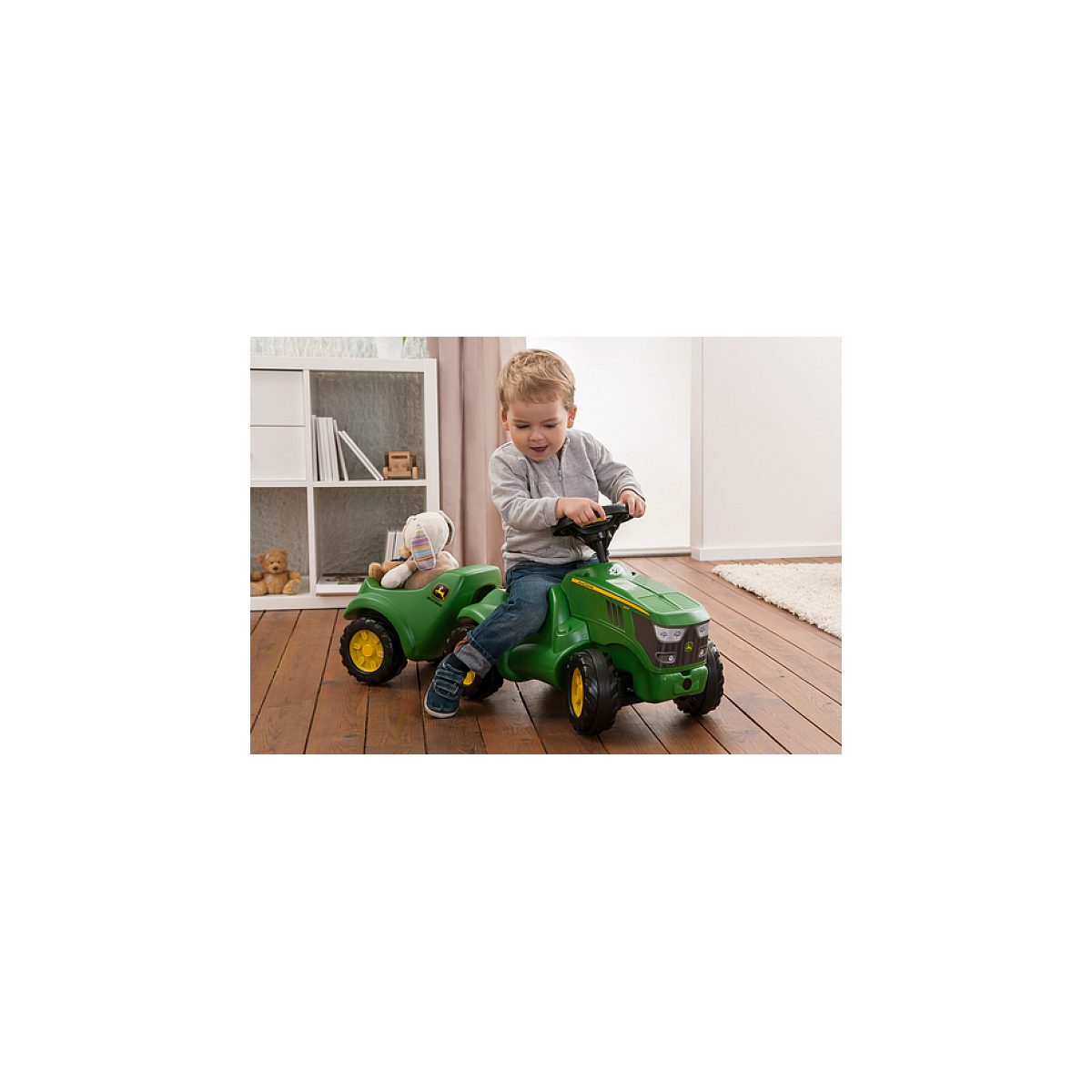Ein kleiner Junge fährt in einem hellen Raum auf einem grünen Spielzeugtraktor mit Stofftieren im Anhänger.