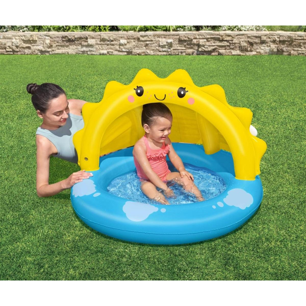 Una mujer se sienta junto a una pequeña piscina inflable azul y redonda con parasol amarillo, donde juega un niño.