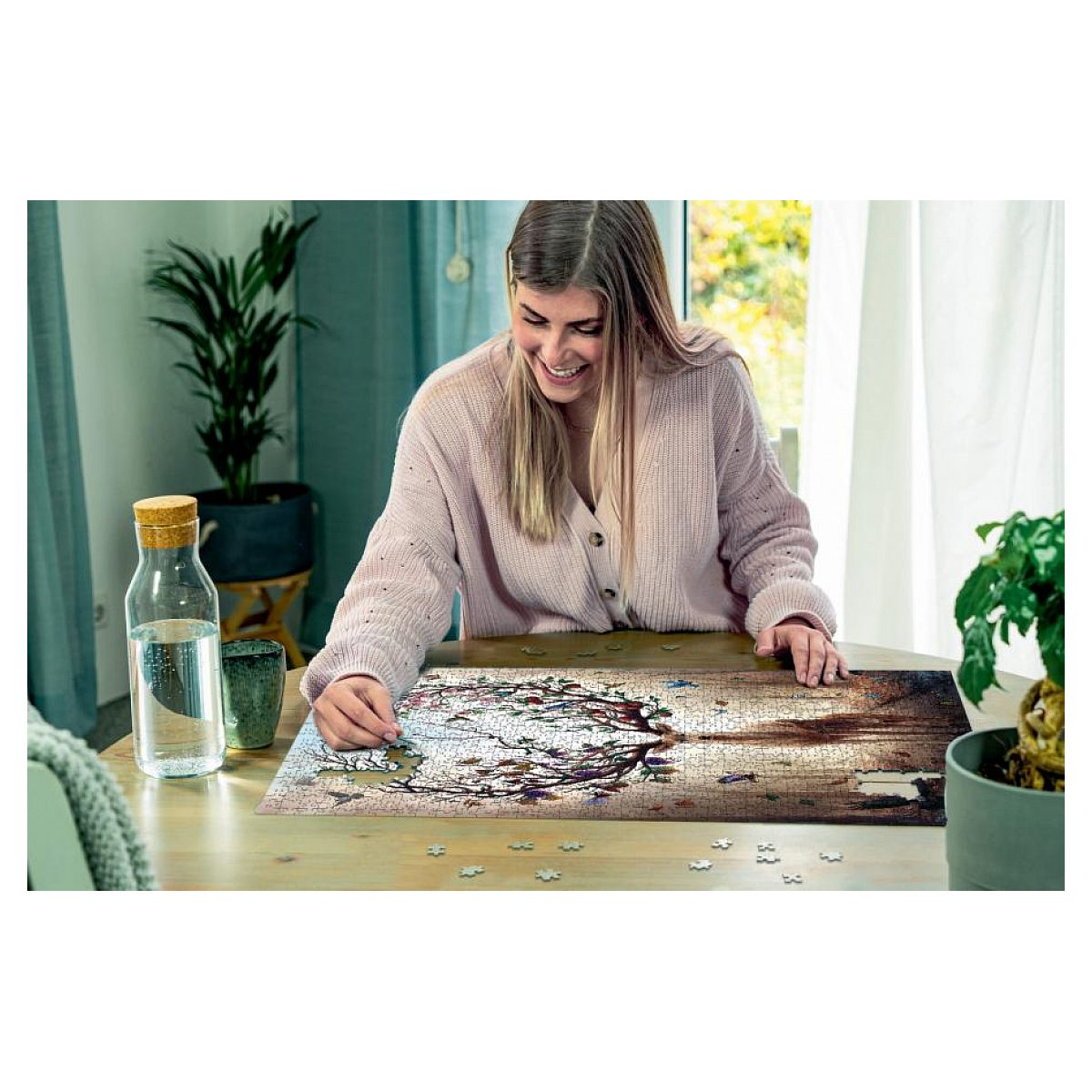 Eine junge Frau sitzt lächelnd an einem Holztisch und setzt ein großes Puzzle mit einem Baummotiv zusammen.