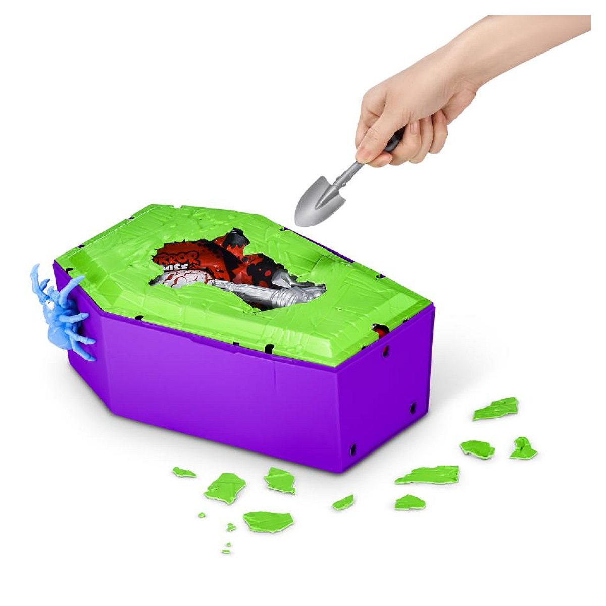 Eine Hand benutzt eine Schaufel, um die grüne Abdeckung einer lila Sargspielzeugbox aufzubrechen.