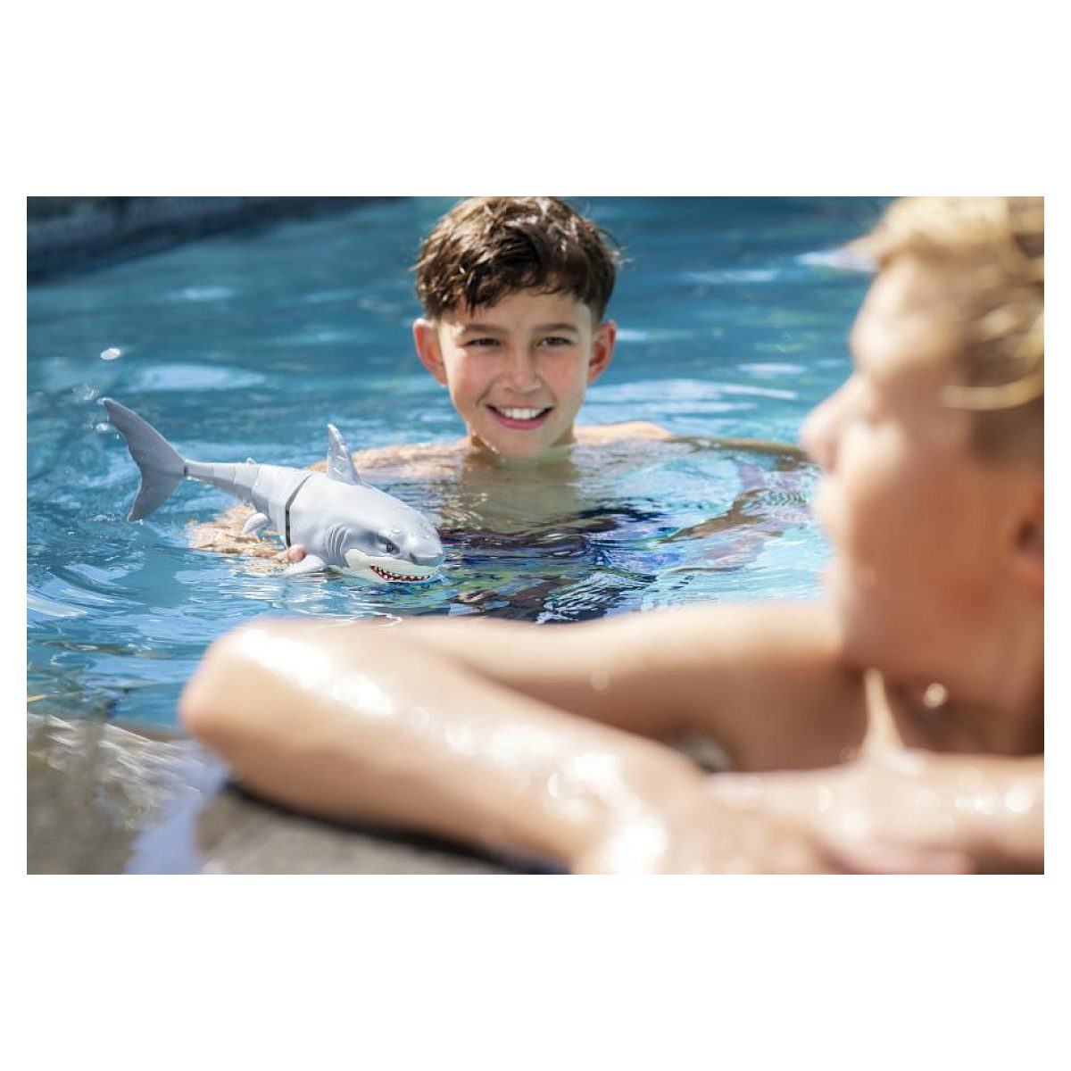 Un niño sonriente en una piscina sostiene un tiburón de juguete, con el brazo de otra persona en primer plano.