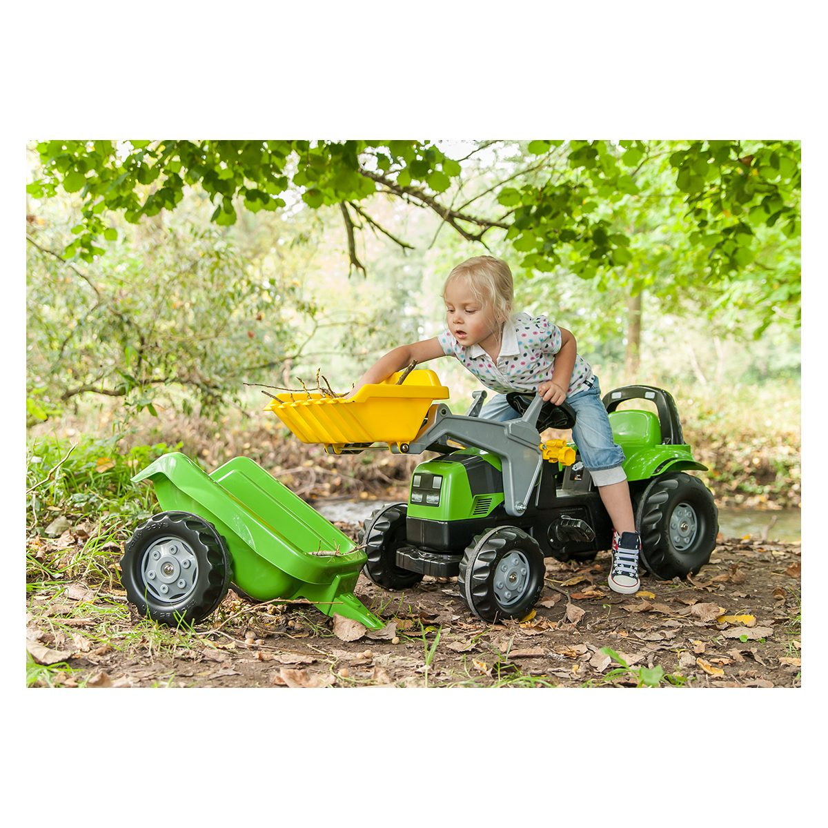 Kleines Kind sitzt im Wald auf einem grünen Spielzeugtraktor mit Anhänger und lädt Zweige in die Schaufel.