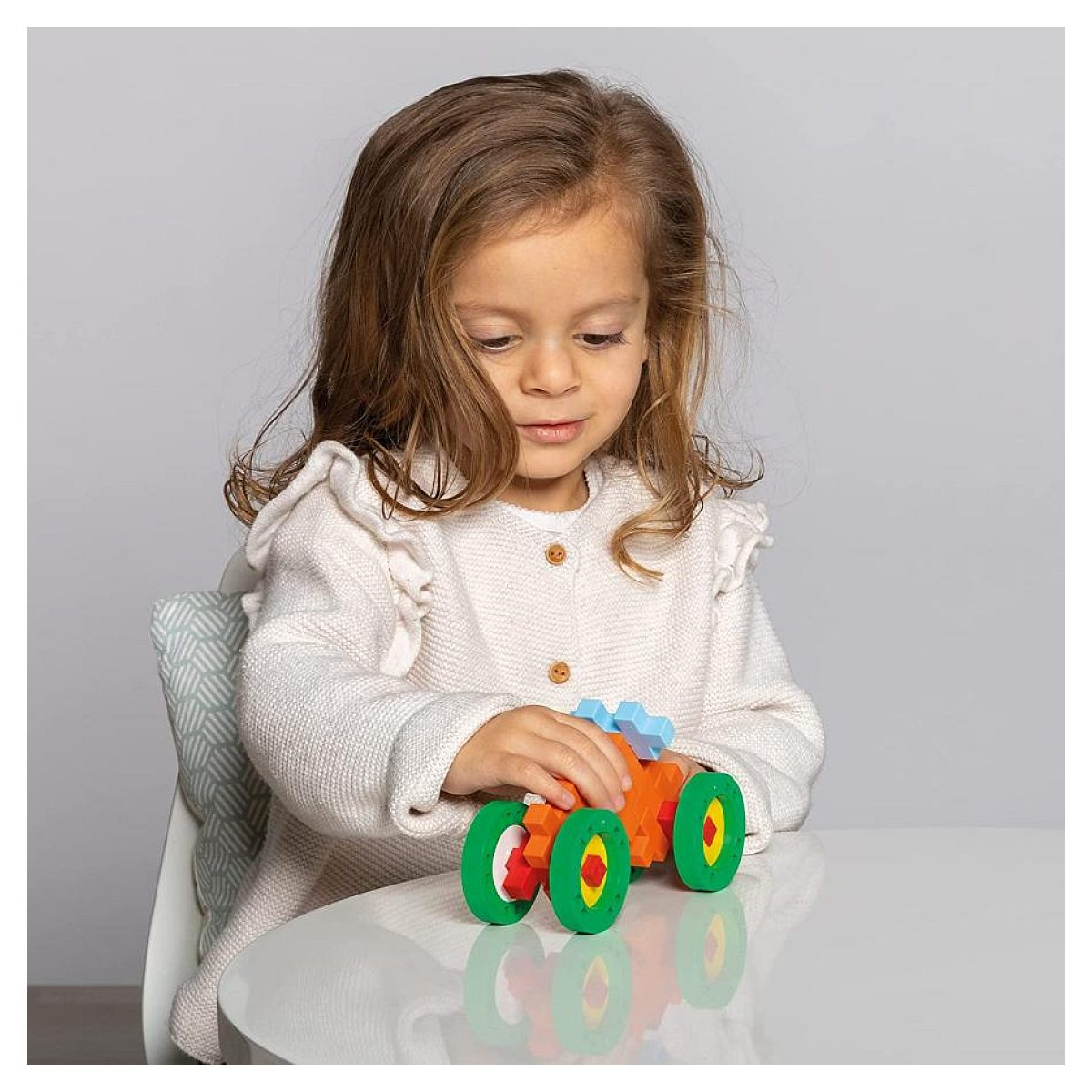 Una bambina siede a un tavolo bianco, concentrata a giocare con una macchina giocattolo colorata.