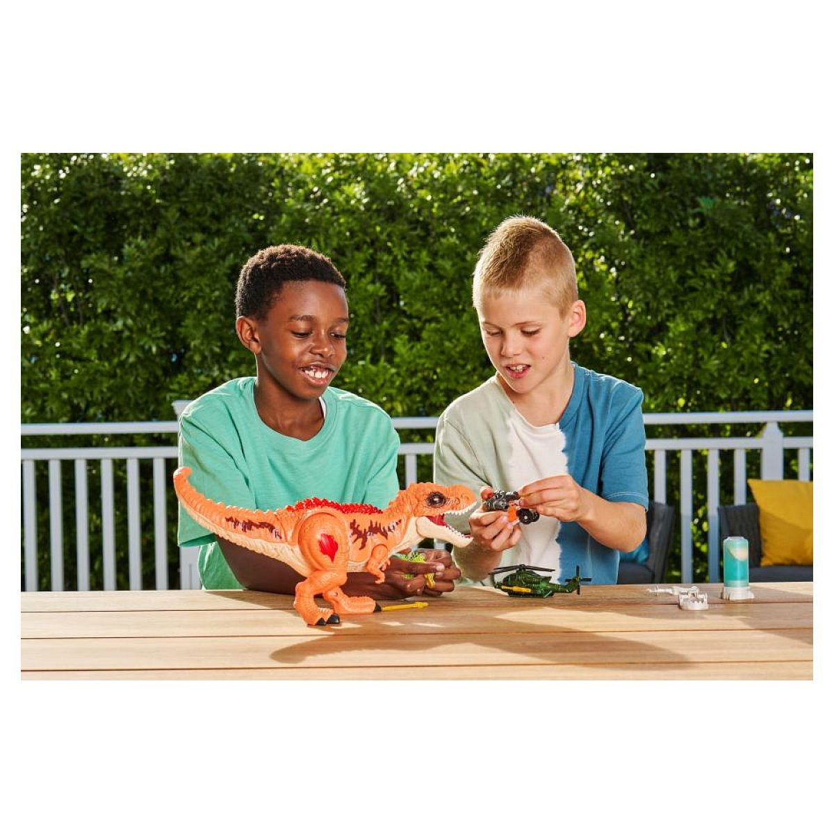 Dos niños juegan al aire libre en una mesa de madera con un gran dinosaurio de juguete naranja y helicópteros.