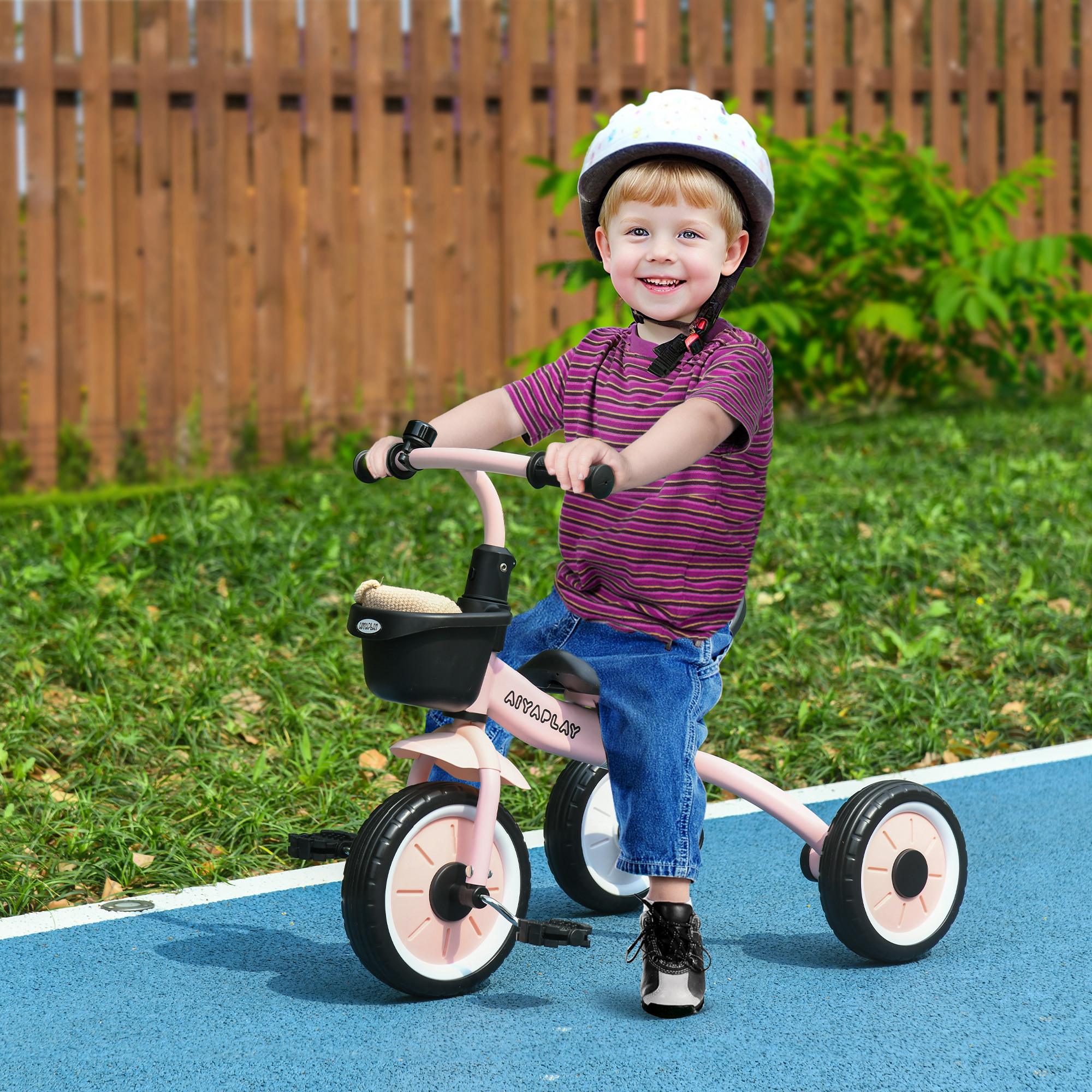 Un bambino sorridente con casco guida un triciclo rosa su un sentiero blu davanti a una staccionata e cespugli.