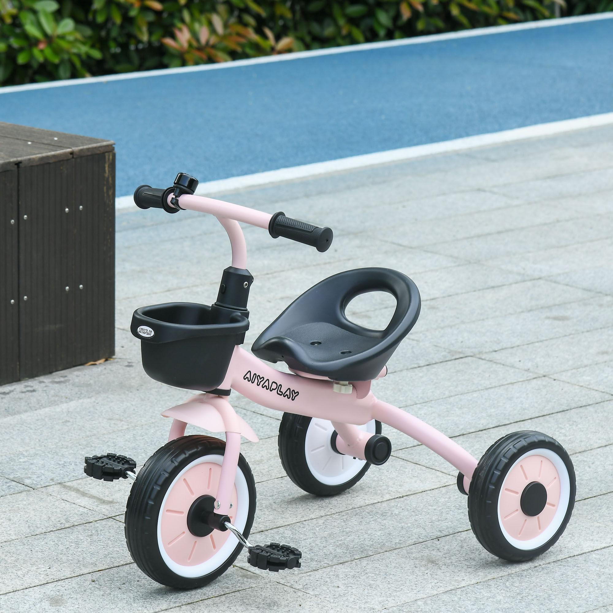Un triciclo per bambini rosa e nero con cestino anteriore su un percorso esterno piastrellato con pista blu sullo sfondo.