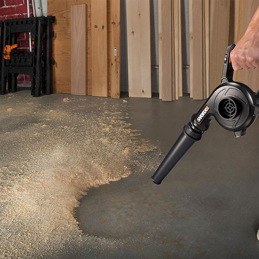 Une main tient un souffleur noir Worx, nettoyant des copeaux de bois sur un sol en béton gris dans un atelier.