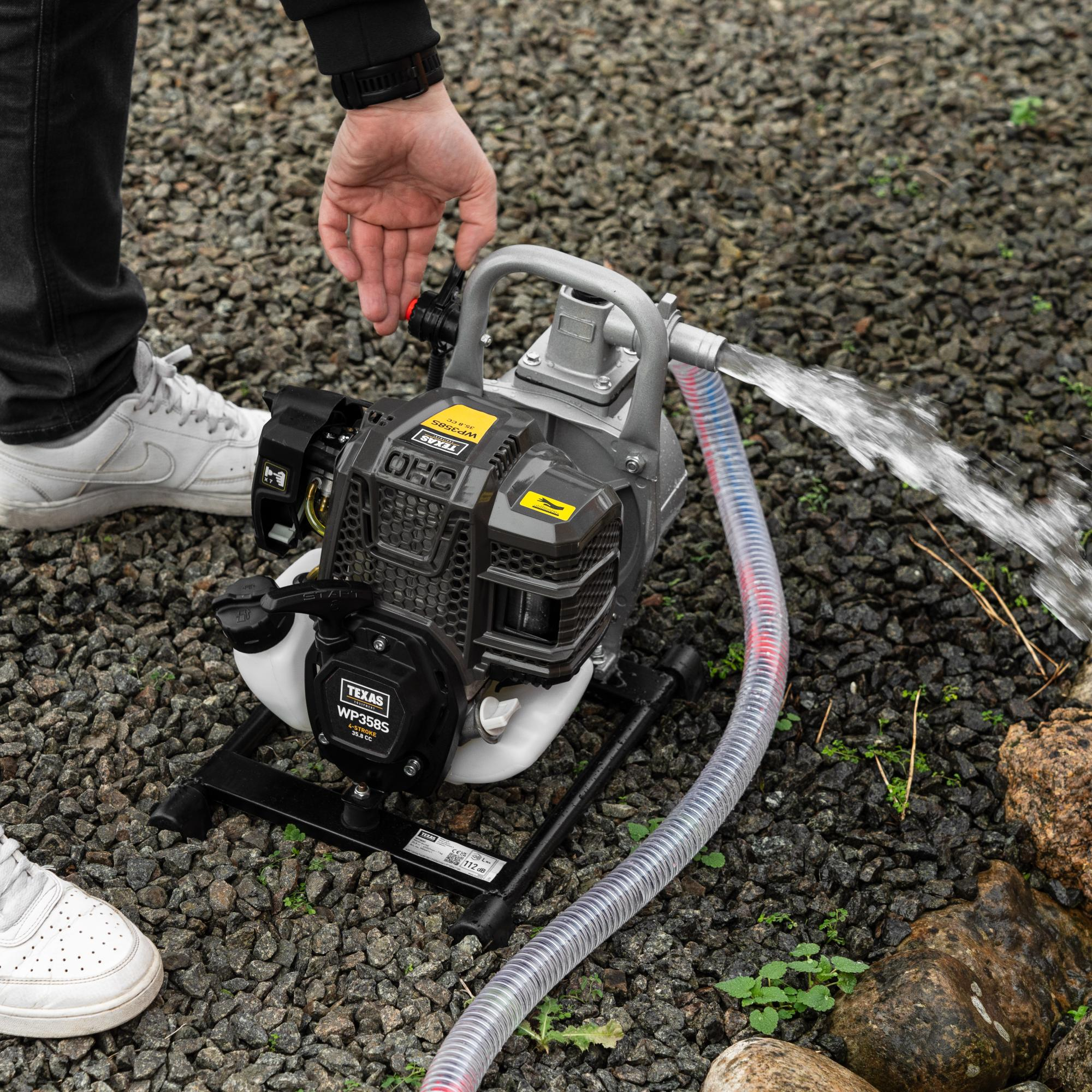 Eine Person startet eine tragbare Benzinwasserpumpe auf kiesigem Boden, während Wasser aus dem Schlauch fließt.