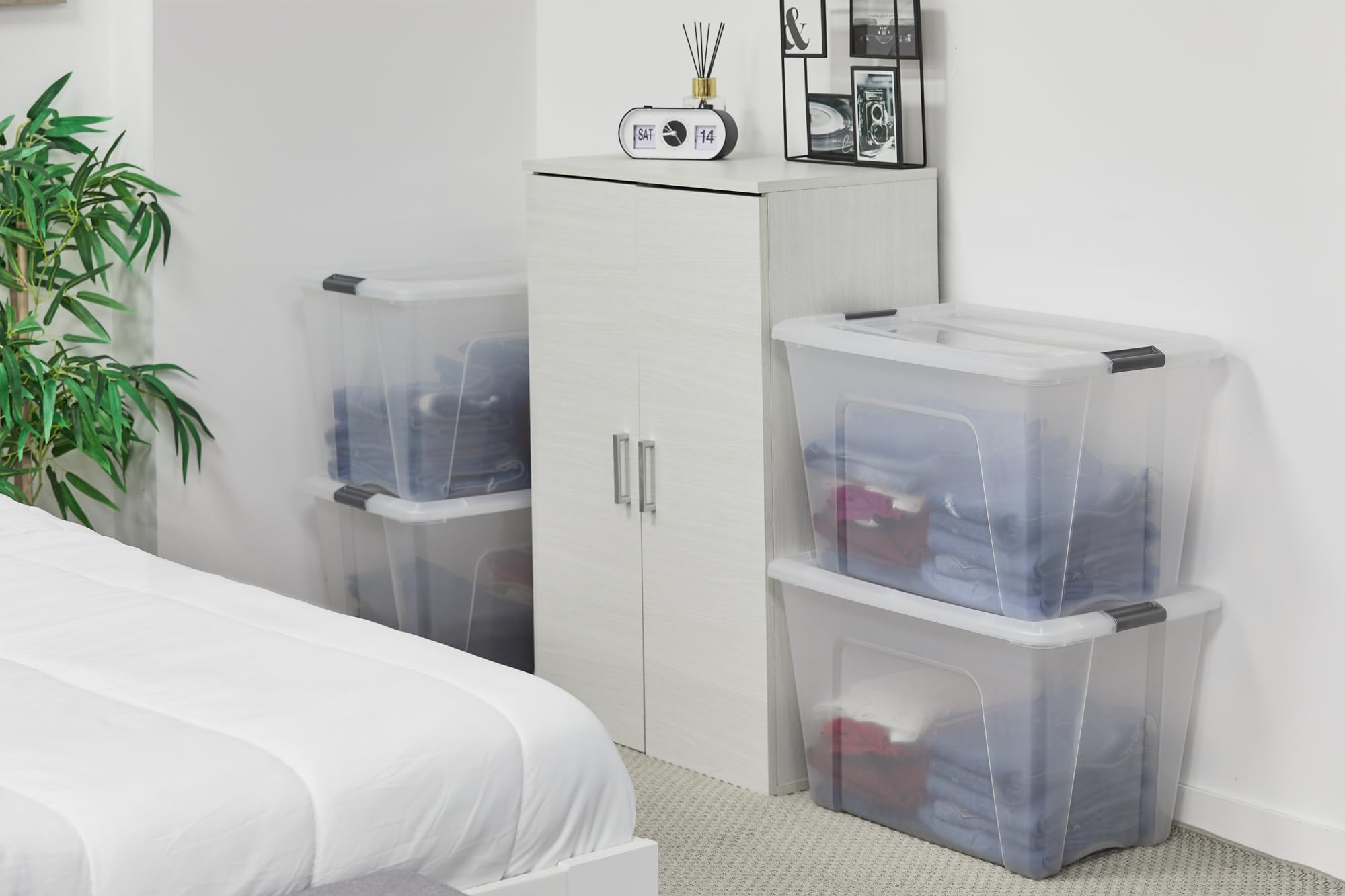 Una camera da letto con scatole di plastica trasparente impilate piene di vestiti vicino a un piccolo mobile bianco.