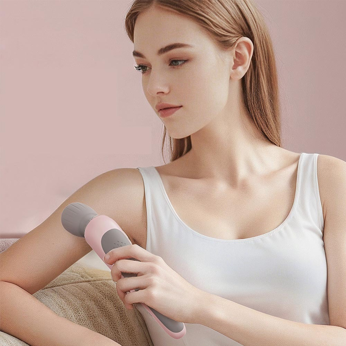 Eine Frau in weißem Tanktop hält ein pink-graues Massagegerät an ihren Arm vor einem pinken Hintergrund.