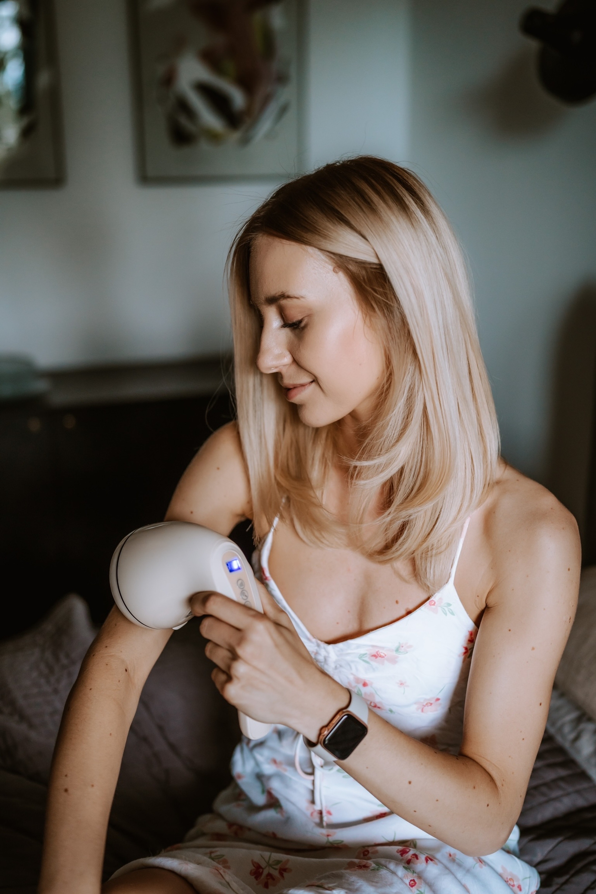 Eine blonde Frau nutzt ein weißes IPL-Haarentfernungsgerät am Arm in einem Schlafzimmer.