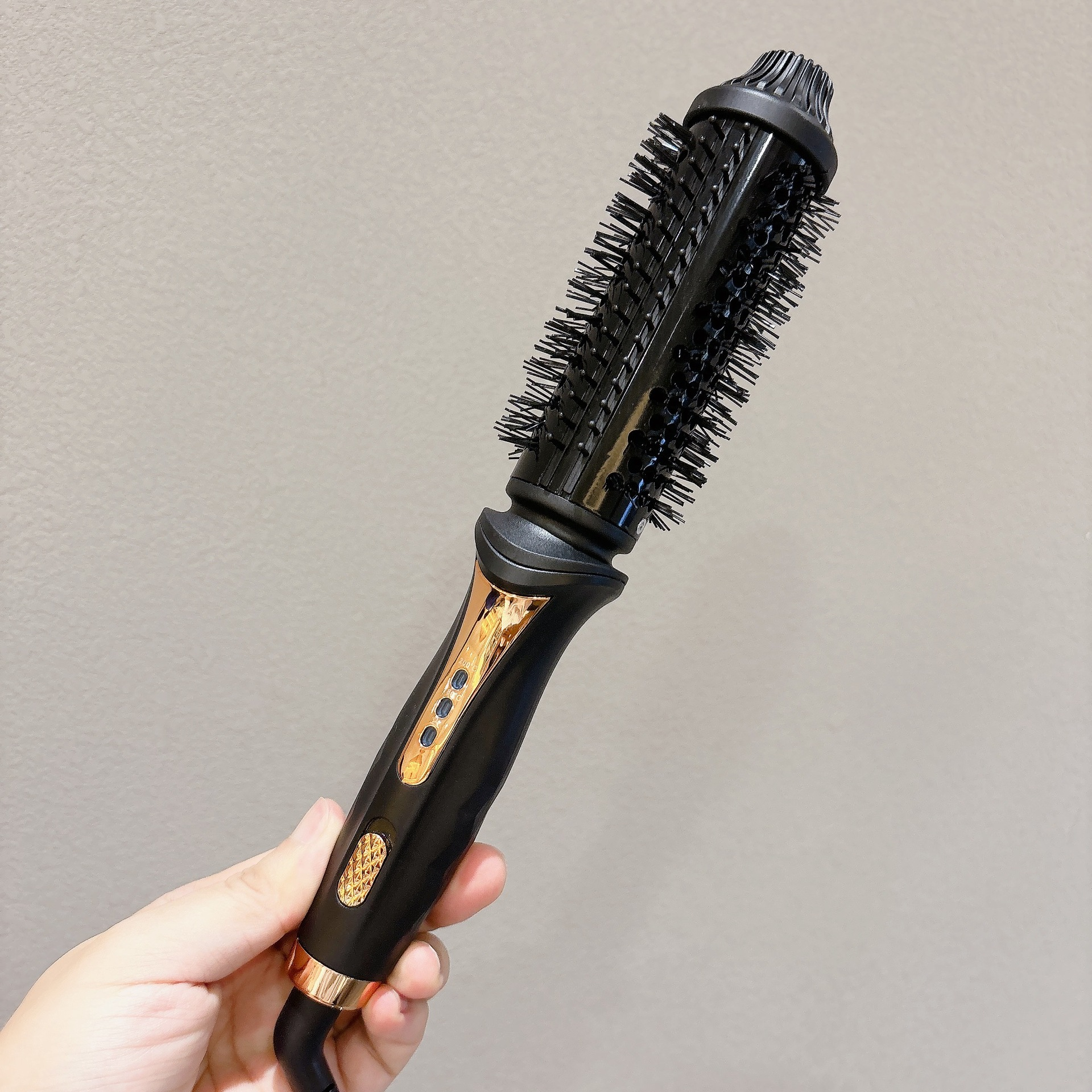 Eine Hand hält eine schwarze Heißluftbürste mit goldenen Akzenten gegen eine helle Wand.
