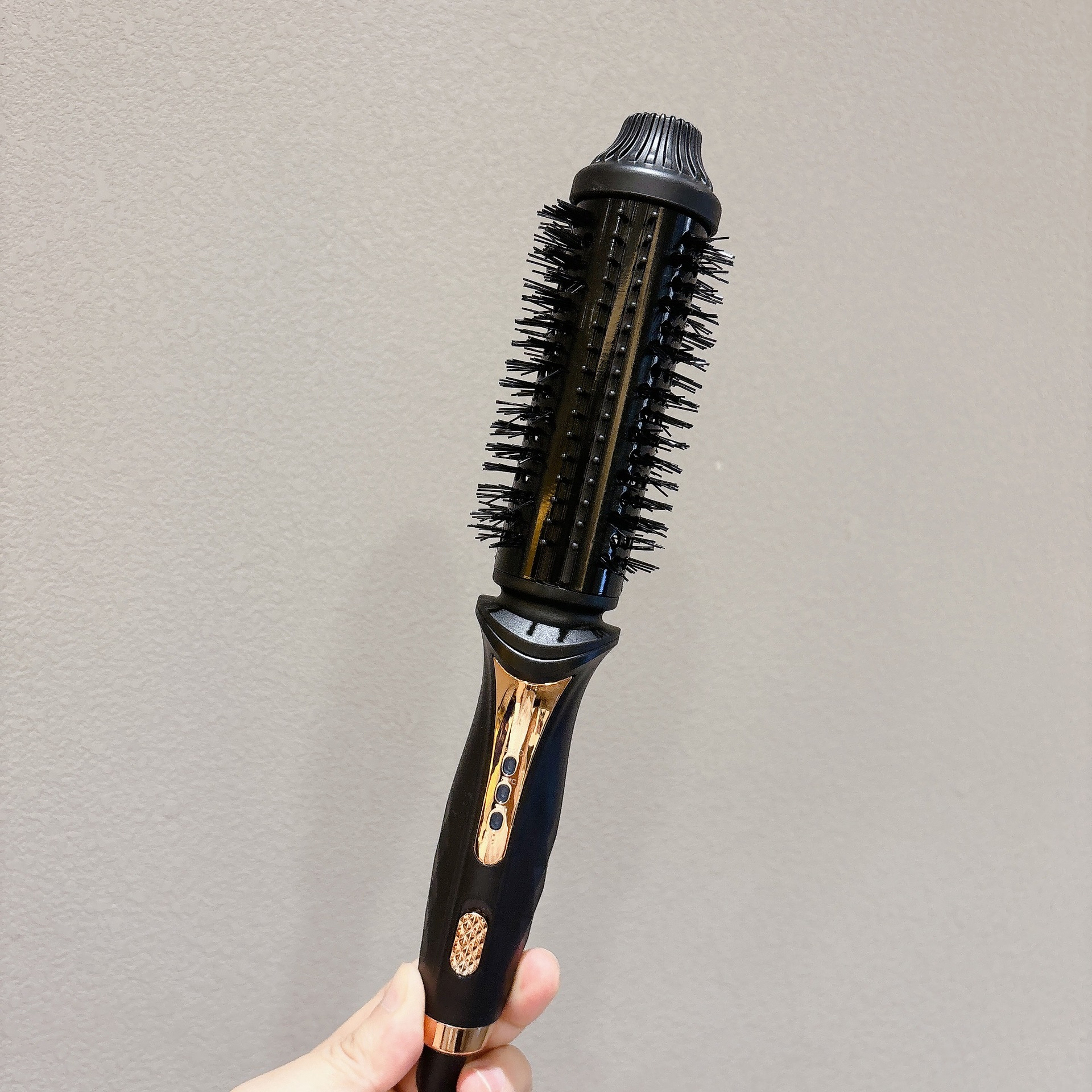 Eine Hand hält eine schwarze und roségoldene elektrische Haarbürste vor einer grauen Wand.