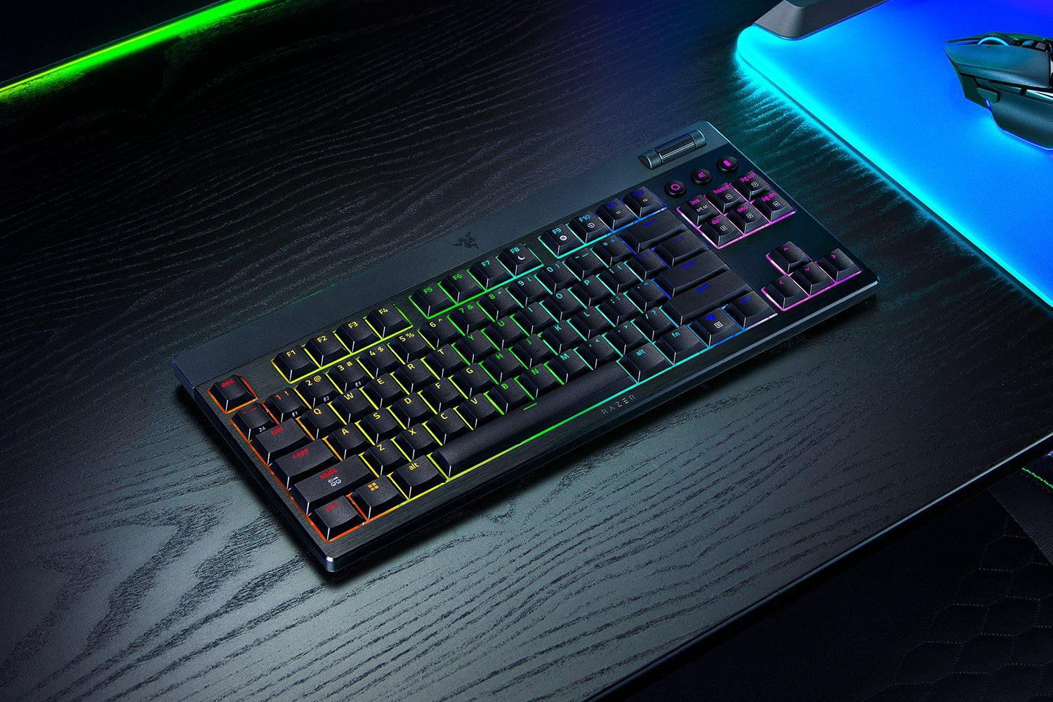 Una tastiera meccanica nera con retroilluminazione RGB su una scrivania in legno accanto a un mouse blu luminoso.