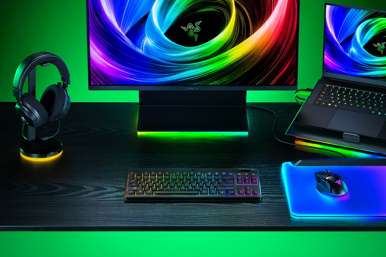 Configurazione di gioco RGB con monitor, laptop, tastiera, mouse su tappetino e cuffie su supporto su sfondo verde.