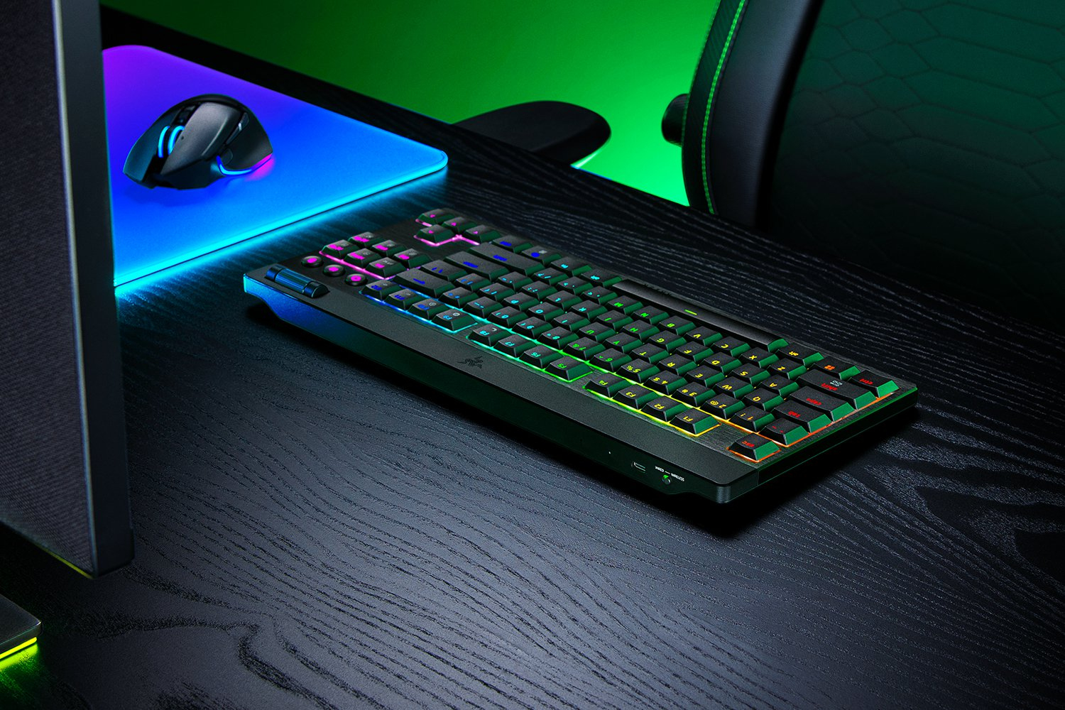 Una tastiera e un mouse RGB illuminati su una scrivania in legno nero con illuminazione ambientale verde e blu.