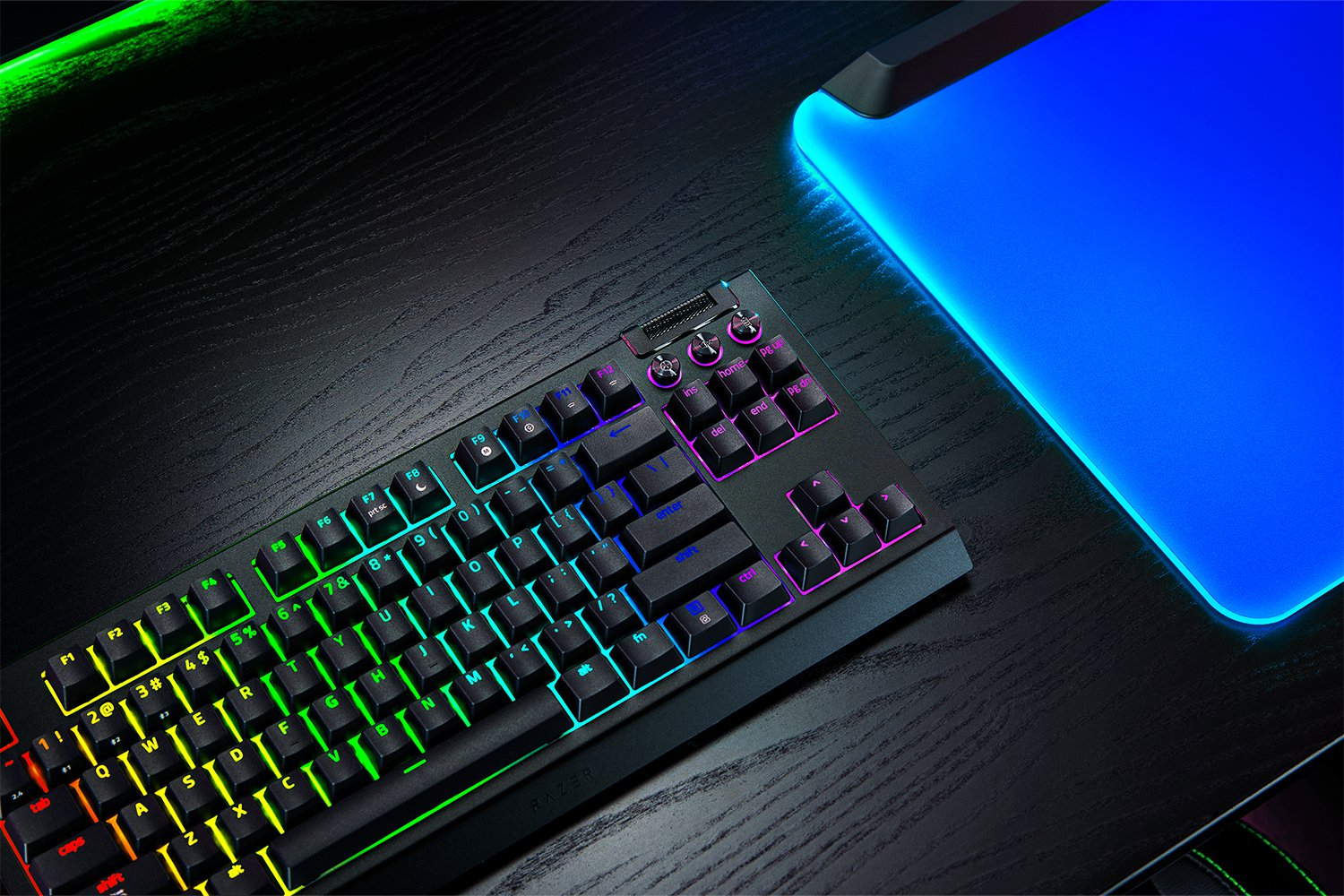 Teclado de juego iluminado y alfombrilla azul en un escritorio de madera oscura.