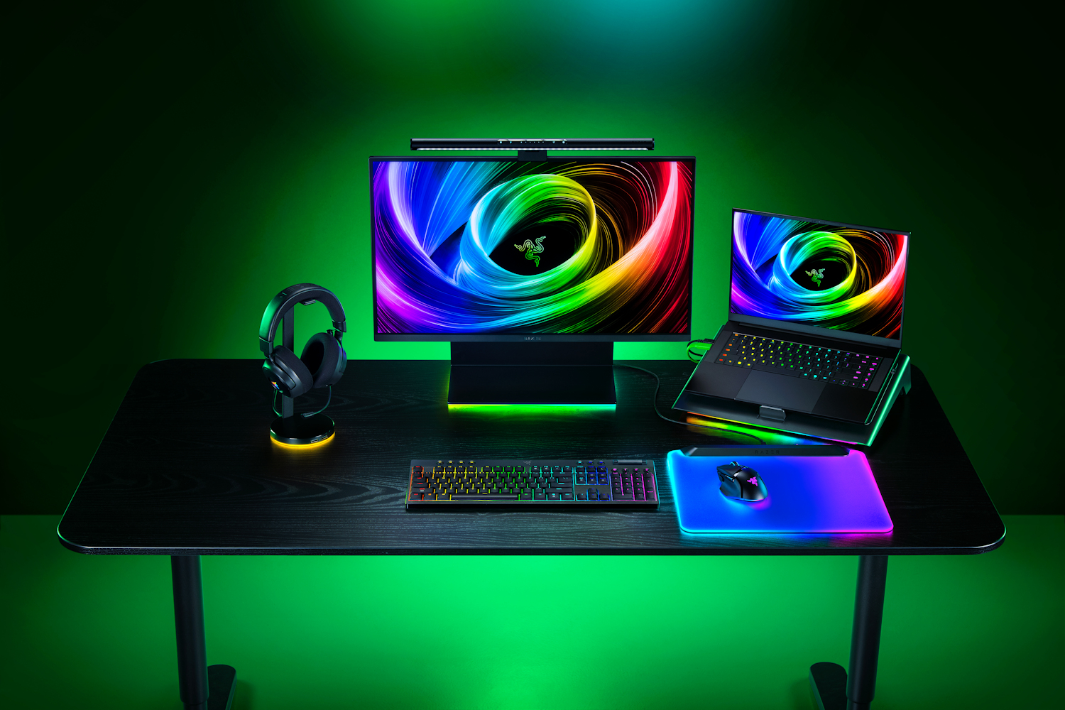 Una scrivania da gioco illuminata con monitor, laptop, tastiera, mouse, cuffie e illuminazione di sfondo verde.