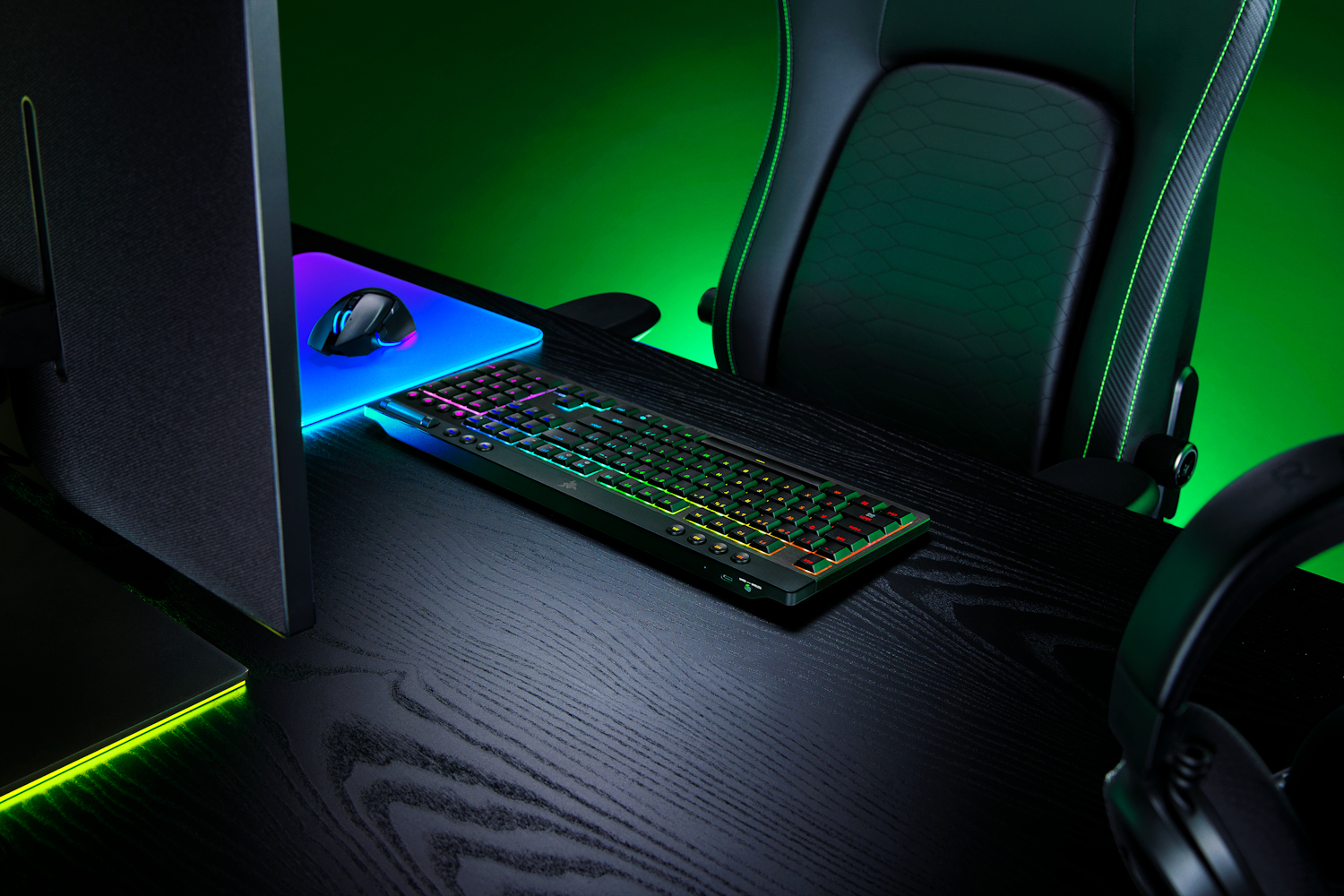 Una scrivania da gaming con tastiera RGB, tappetino luminoso, mouse e cuffie su sfondo verde.