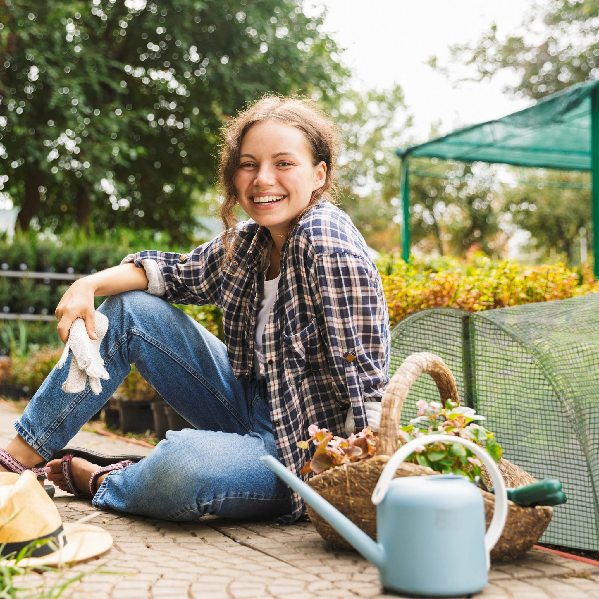 Eine lächelnde junge Frau sitzt in einem Garten mit einer blauen Gießkanne und Pflanzen.