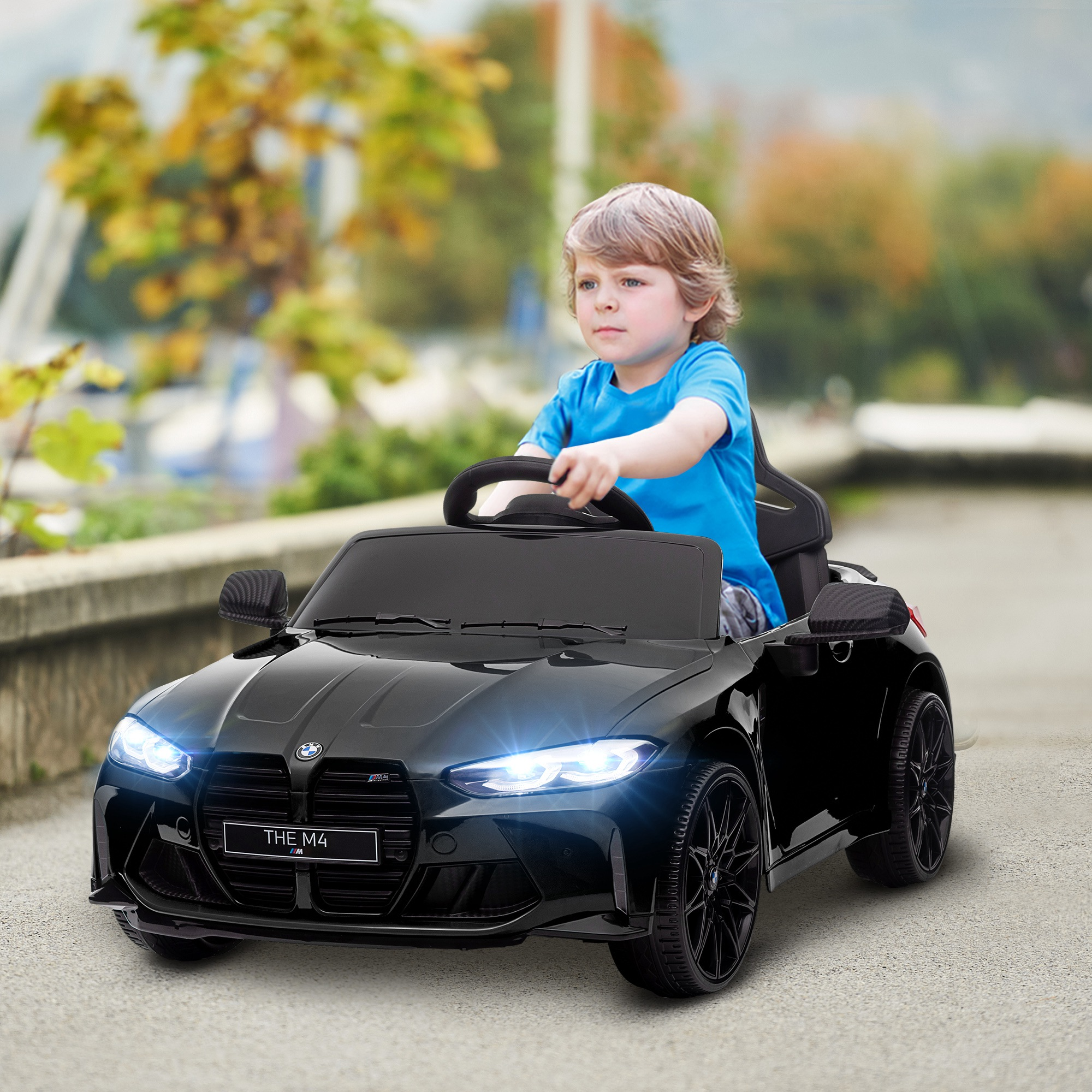 Un bambino biondo con una maglietta blu guida un'auto BMW giocattolo nera all'aperto con i fari accesi.