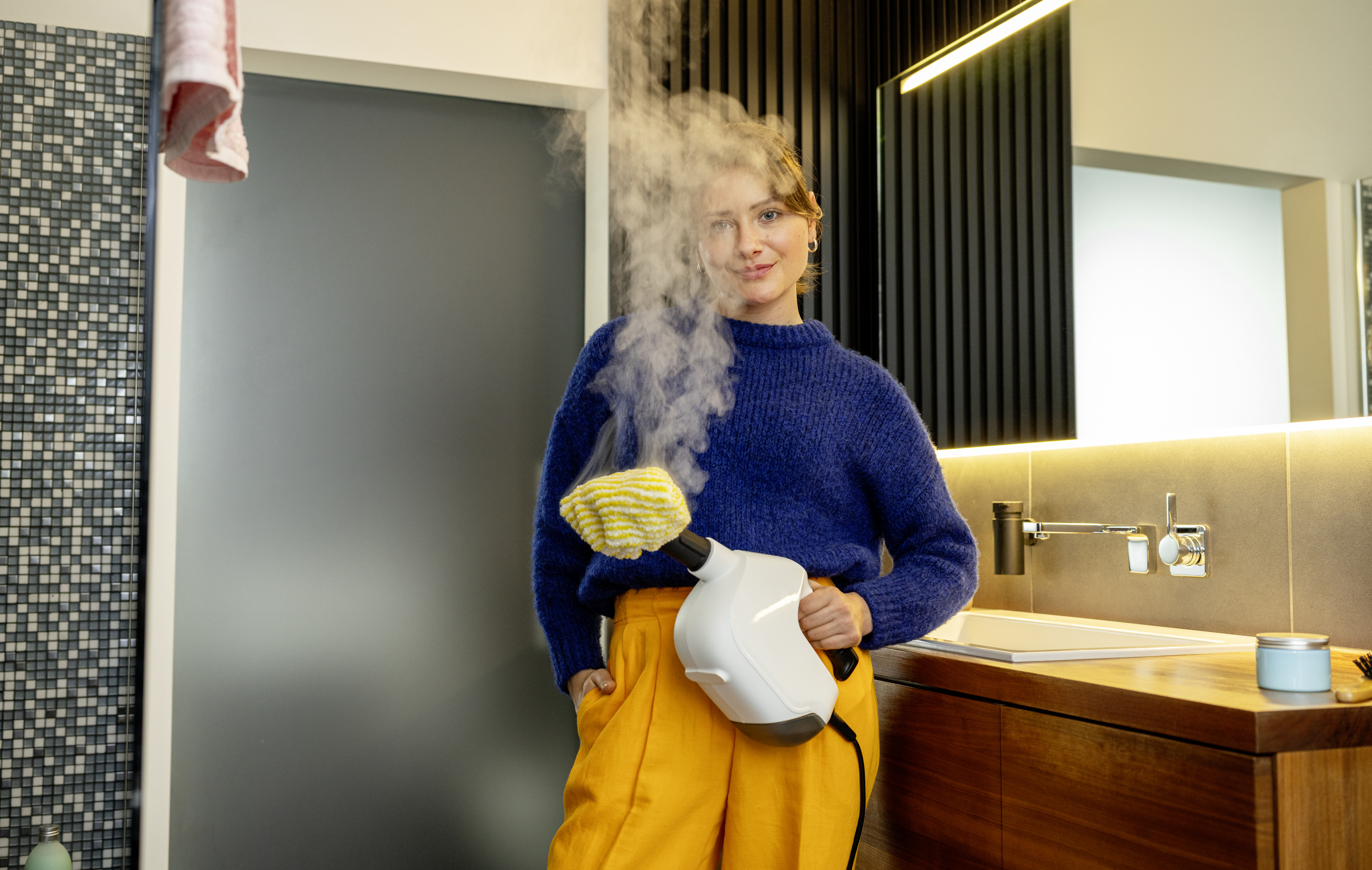 Eine Frau in blauem Pullover und gelber Hose hält einen Dampfreiniger in einem modernen Badezimmer.