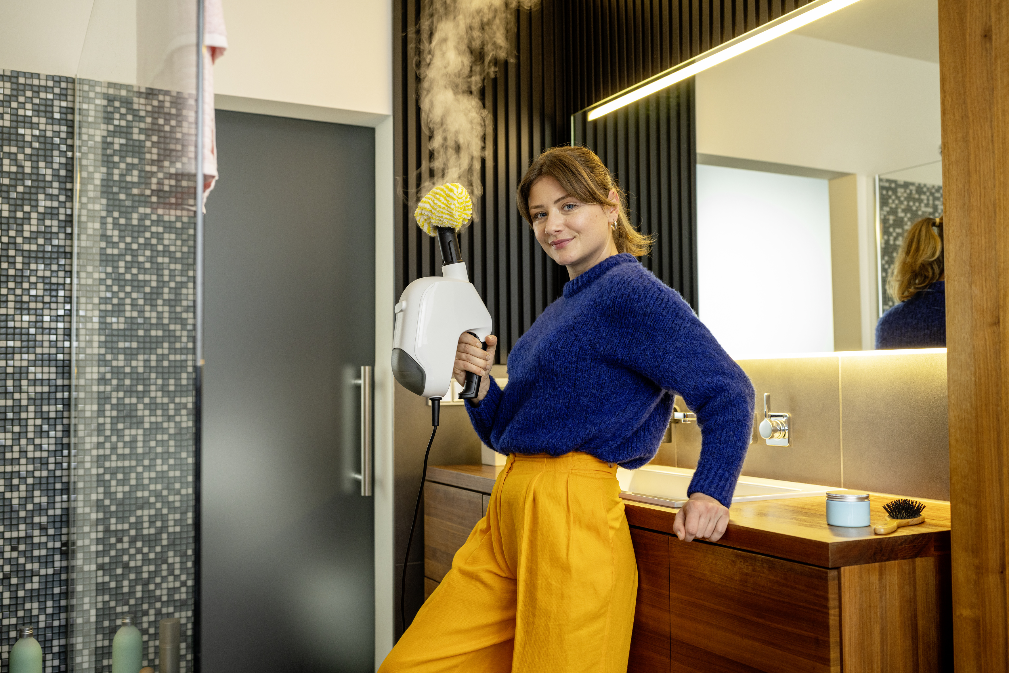 Eine Frau in blauem Pullover und orangefarbenen Hose hält einen dampfenden Reiniger im modernen Badezimmer.