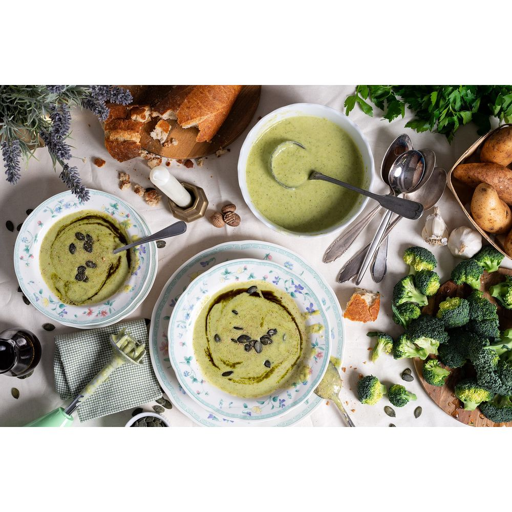 Zuppa verde in ciotole su una tavola apparecchiata con pane, broccoli, patate e posate.